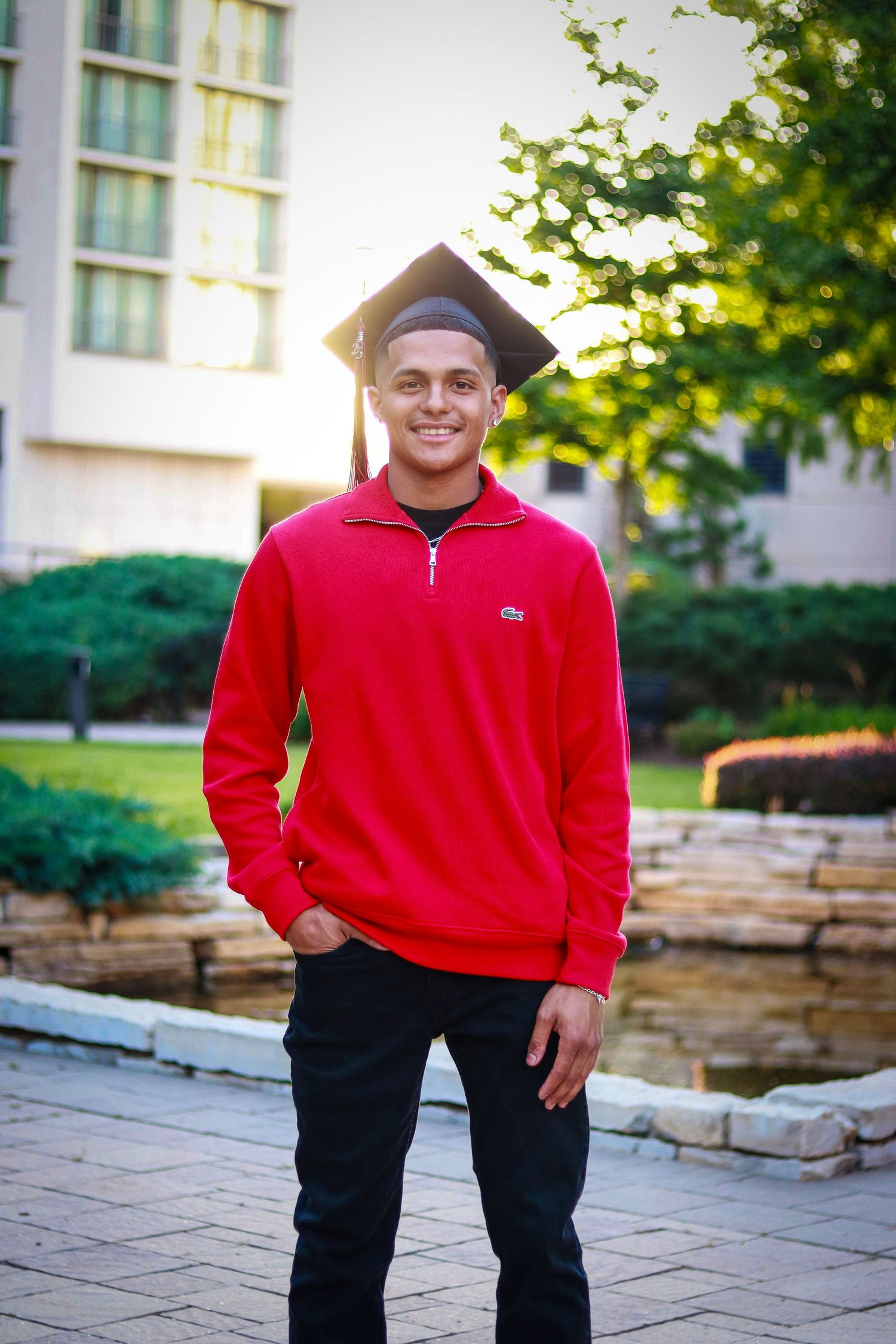 A person in a red fleece pullover and dark pants stands outdoors on a stone patio near a building.