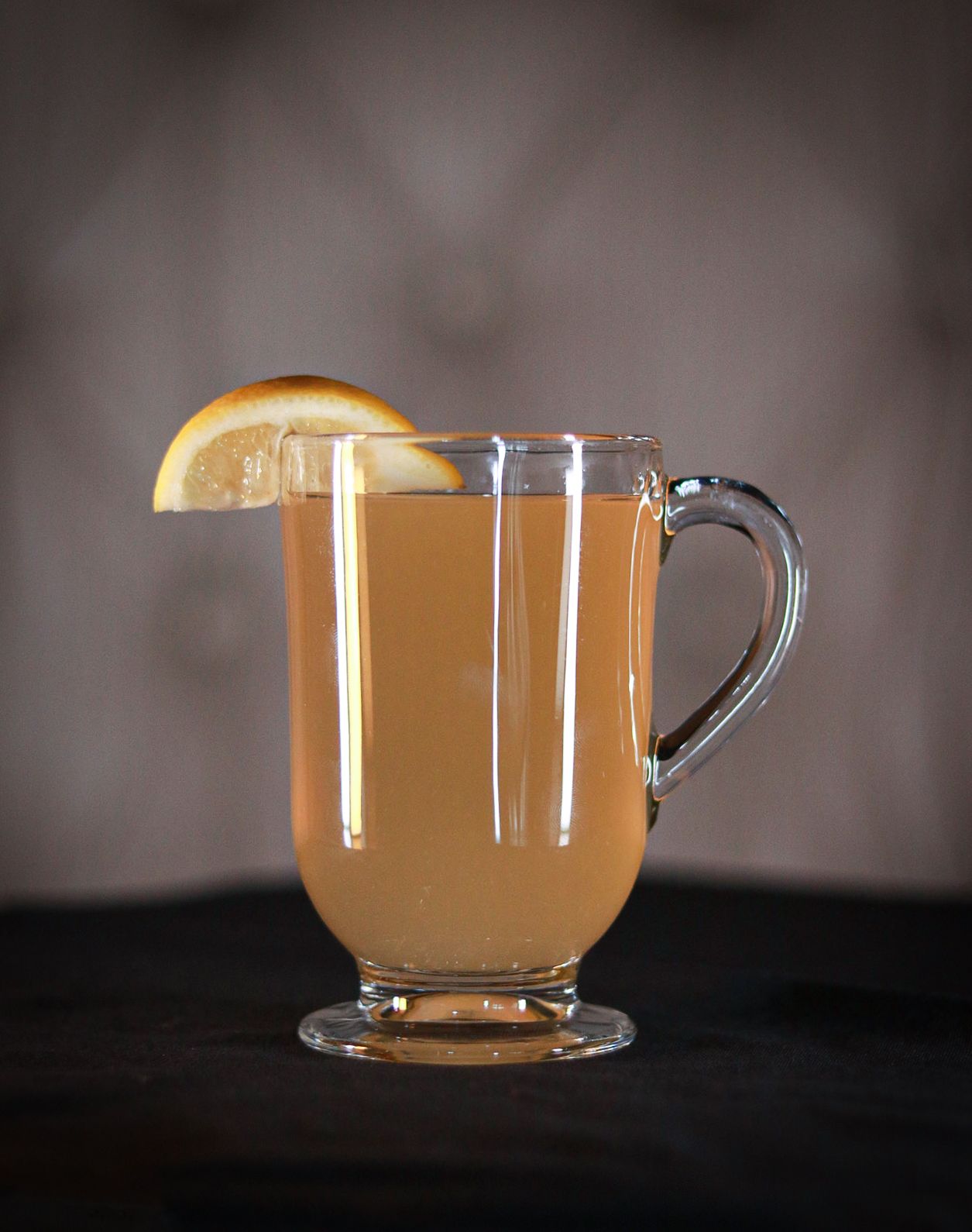 Hot beverage with orange slice garnish served in clear glass mug against moody backdrop.