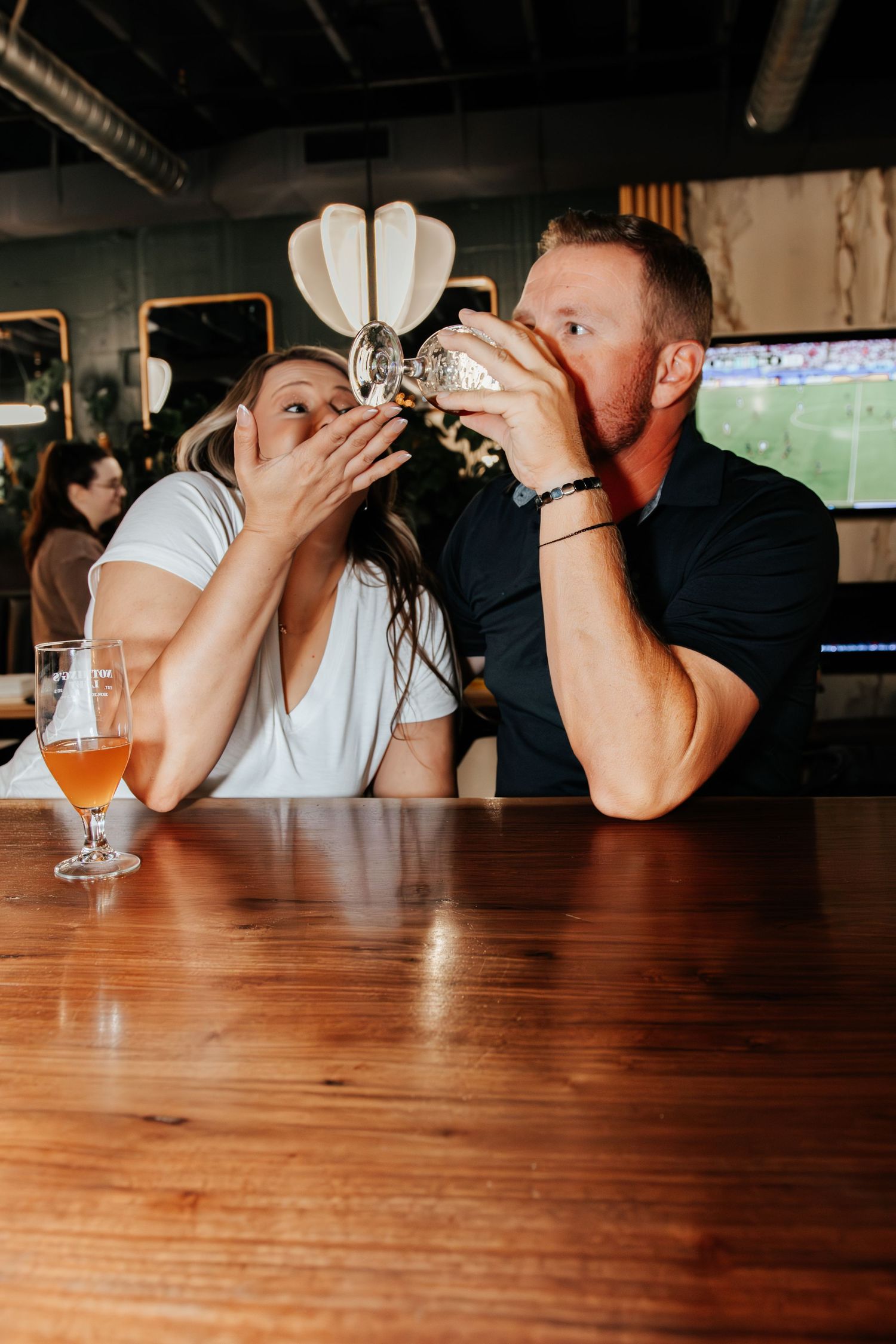 Couple sharing a drink together at a bar, with a glass of craft beer on the wooden counter and a TV in the background.