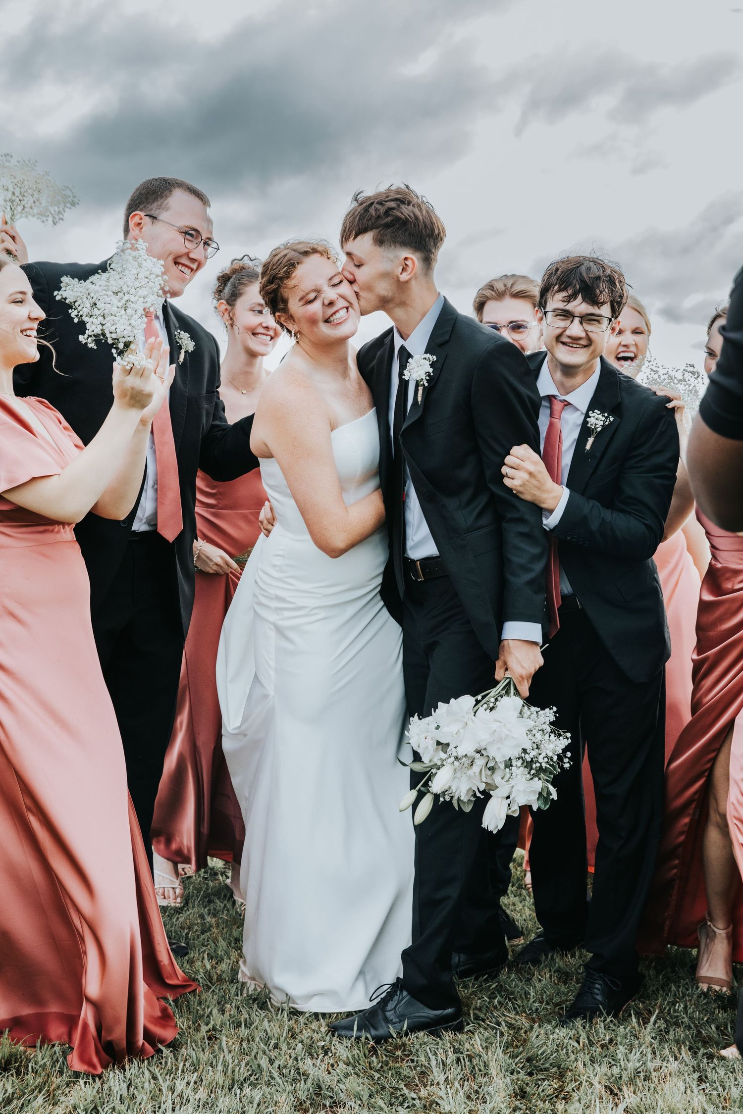 Bride and groom share a kiss surrounded by smiling wedding party in coral dresses and black suits outdoors.