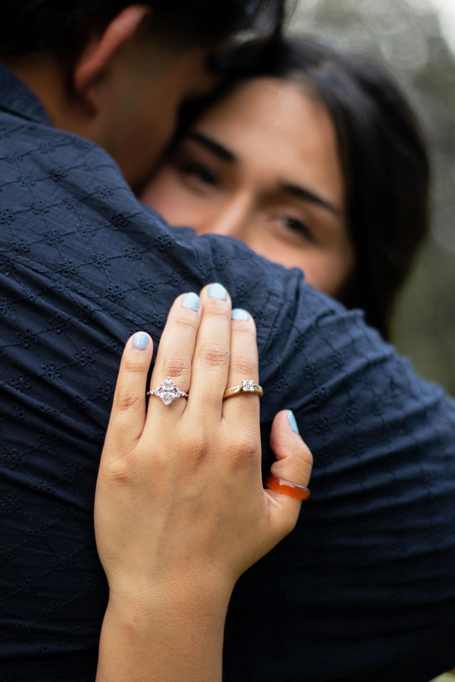 Close-up of woman's hand displaying a diamond engagement ring while embracing her fiance outdoors.
