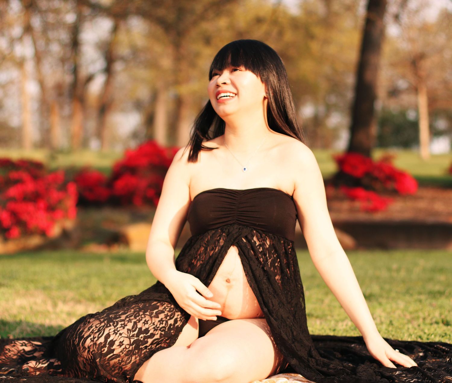 A person in black lace dress poses outdoors in a park setting with autumn trees and red flowers in the background.