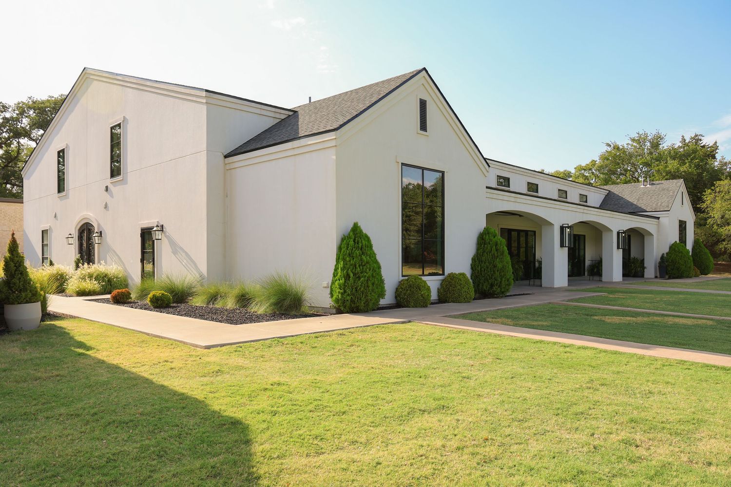 Modern white farmhouse exterior with arched portico, manicured shrubs, and lush green lawn on a sunny day.