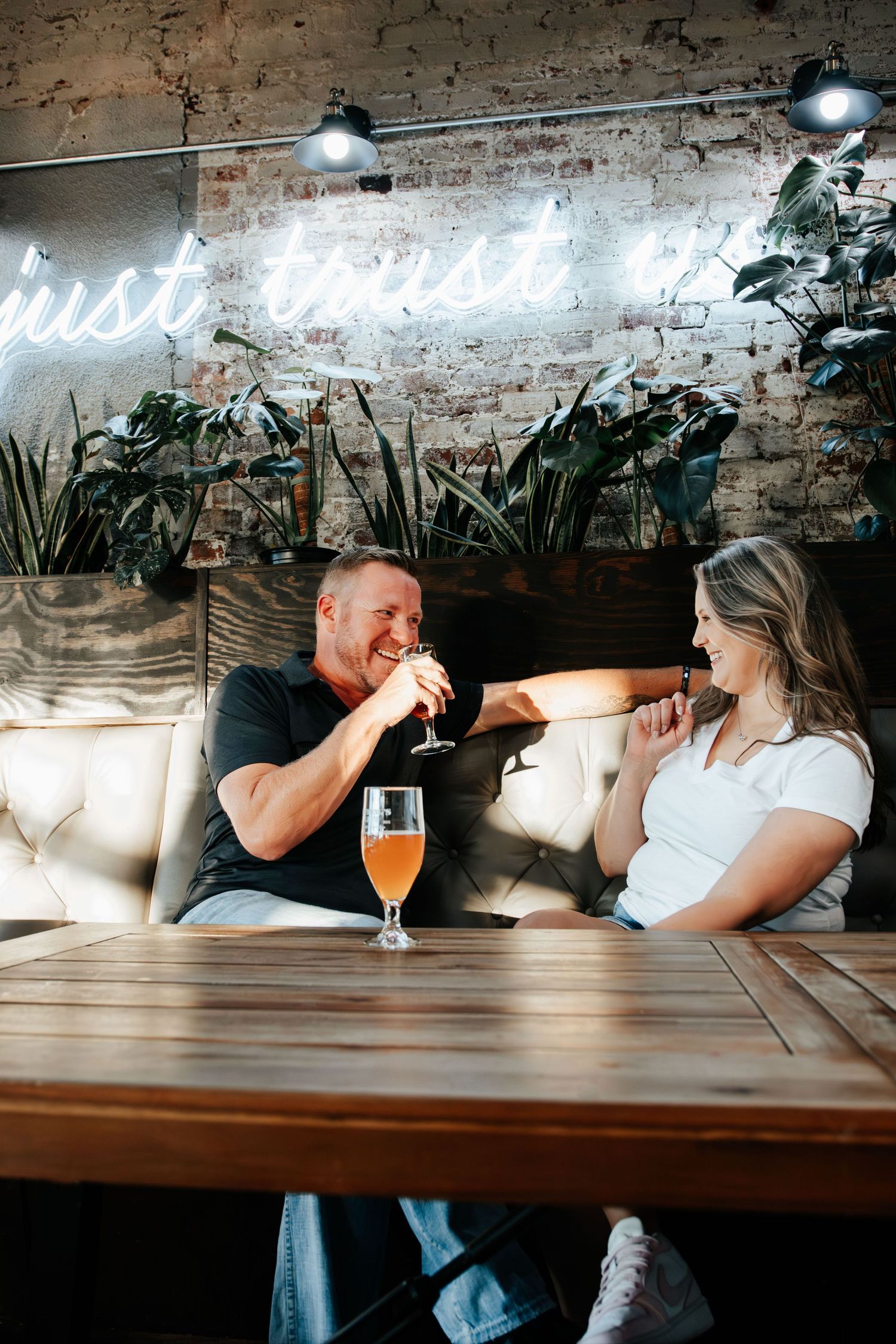 Couple laughing and clinking glasses of craft beer at a rustic bar with neon sign and exposed brick wall.