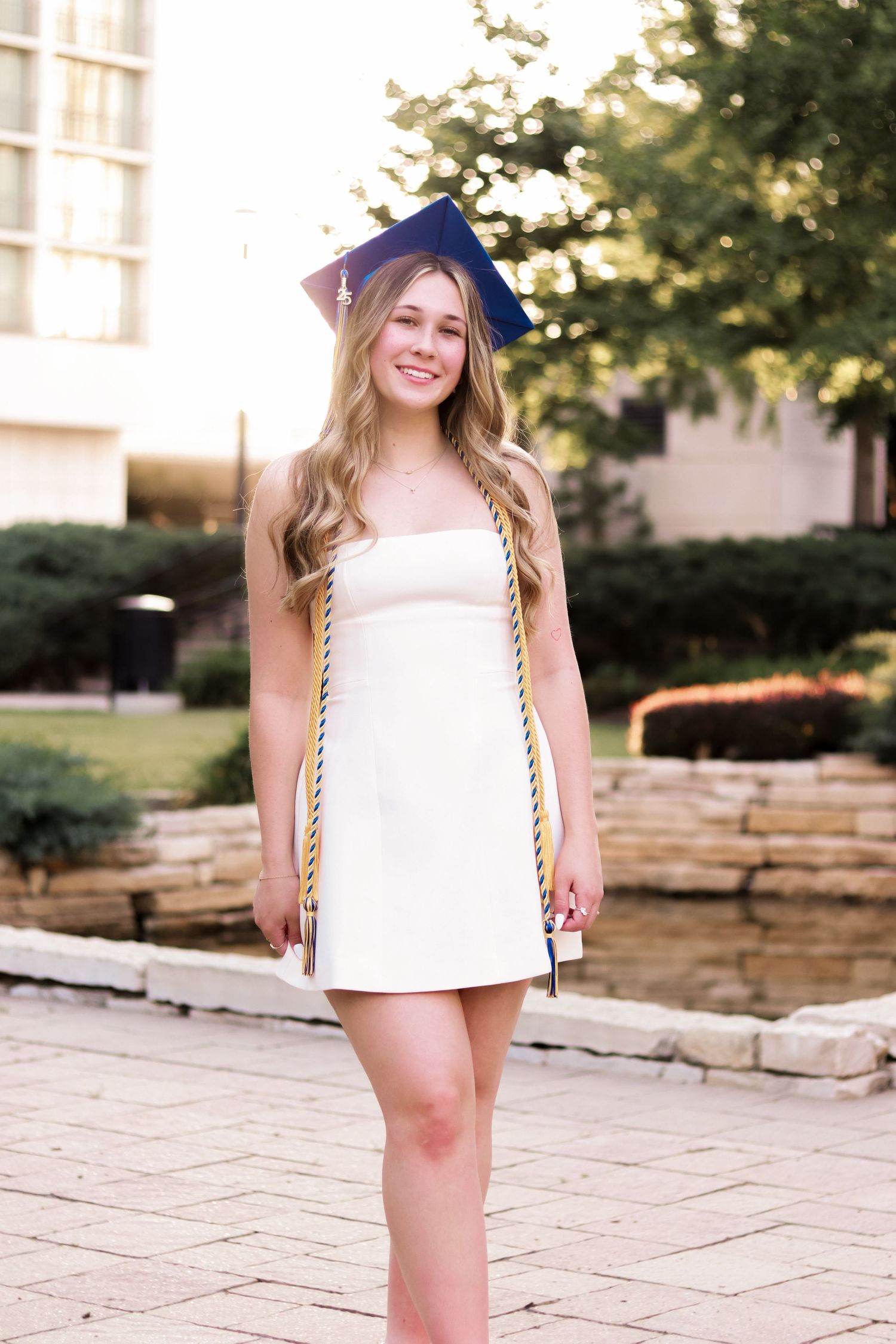 Graduate in white dress and blue cap with honor cords stands on stone patio during outdoor photoshoot.