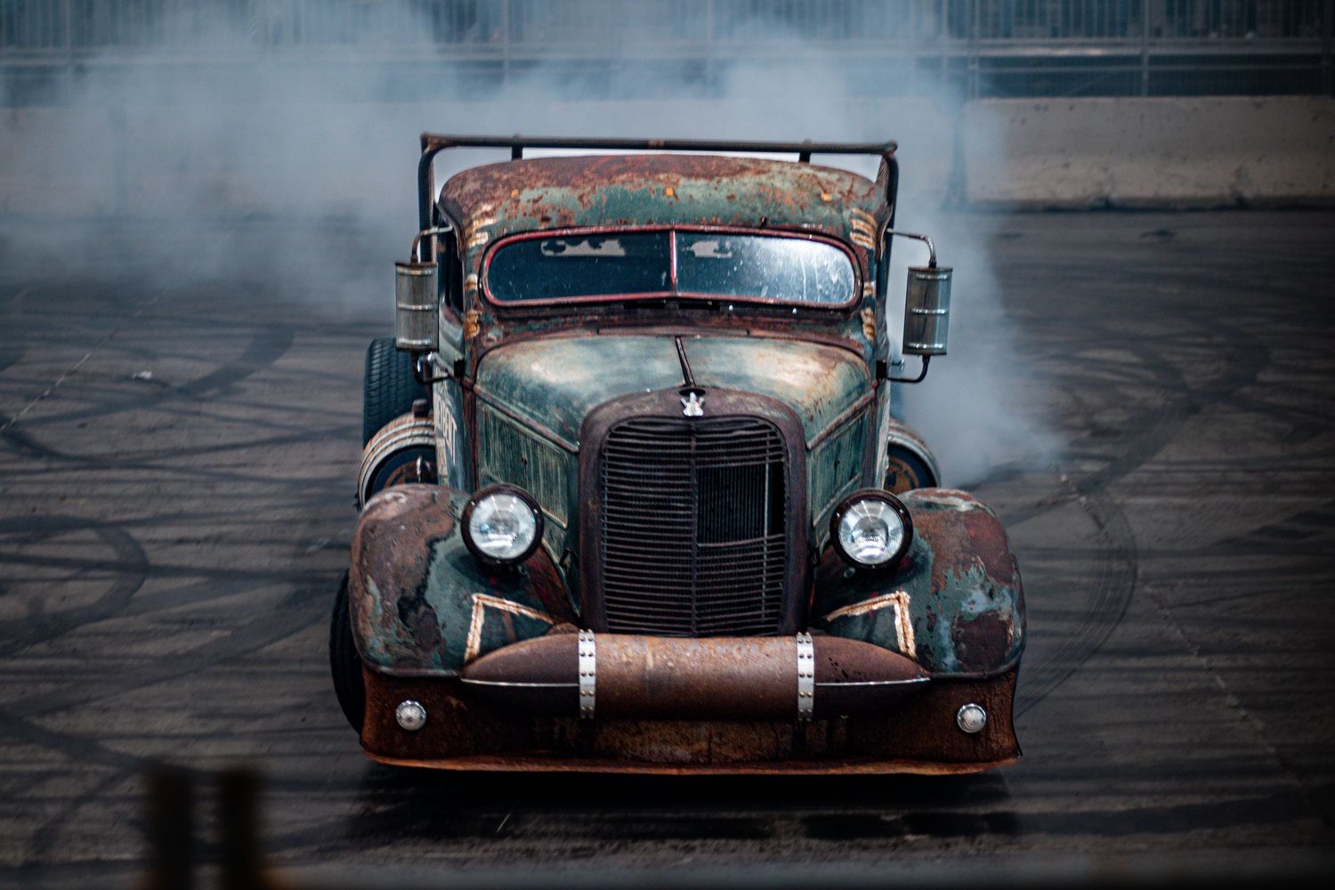 Vintage rusty rat rod truck performing a burnout, releasing thick white smoke on a dark asphalt surface.
