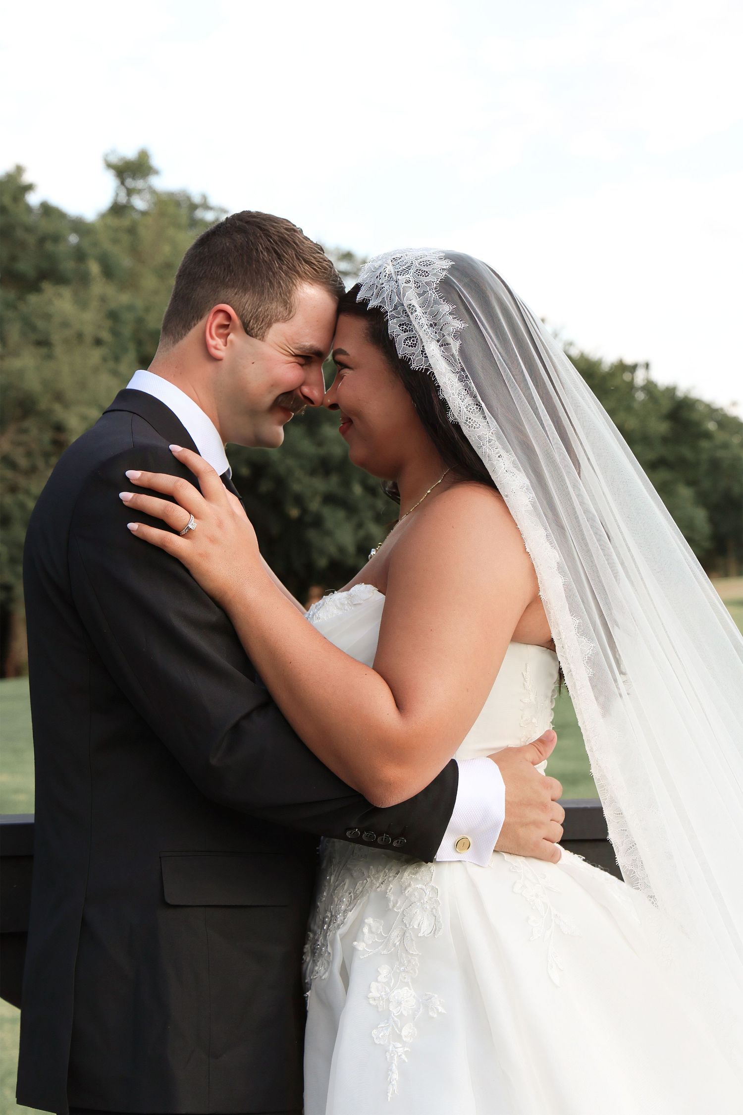 Bride and groom embrace tenderly in wedding attire, sharing an intimate moment outdoors with lush green trees in background.