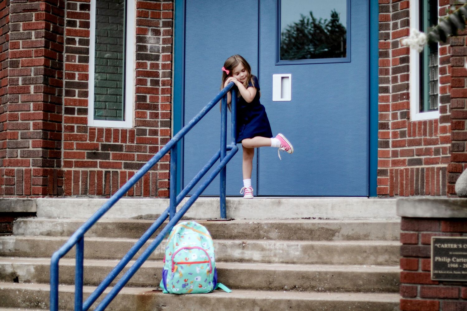 A blue backpack sits on concrete steps outside a brick building with blue railings and white trim.