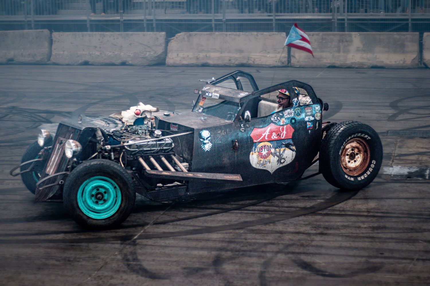A custom rat rod race car with teal wheels and stickers speeds on a track with a Puerto Rican flag in the background.