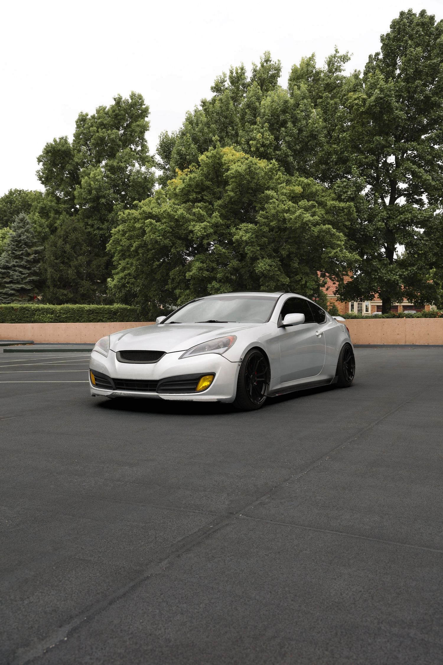 Silver Hyundai Genesis Coupe with yellow accents parked in an empty lot surrounded by lush green trees.