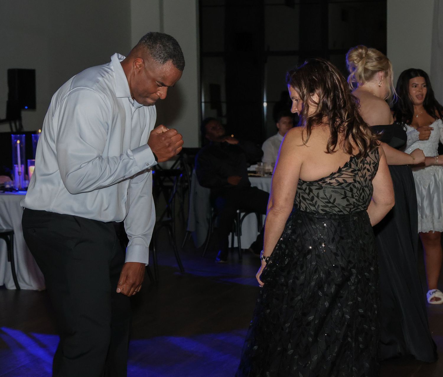 Couple dancing together at a formal event, man in white shirt and woman in black dress, with guests seated in background.