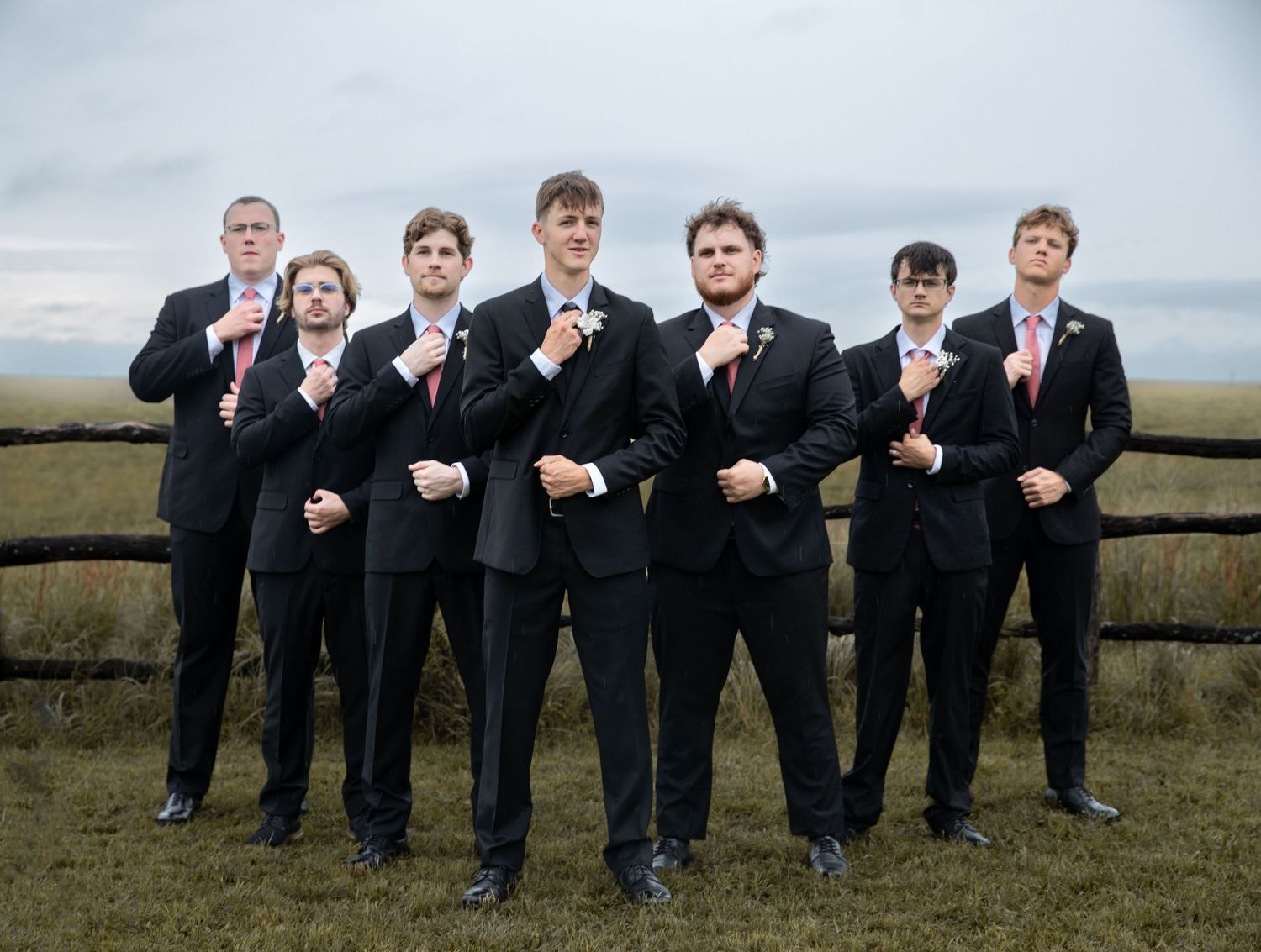 Groomsmen in black suits and red ties posing outdoors near a wooden fence with open fields in the background.