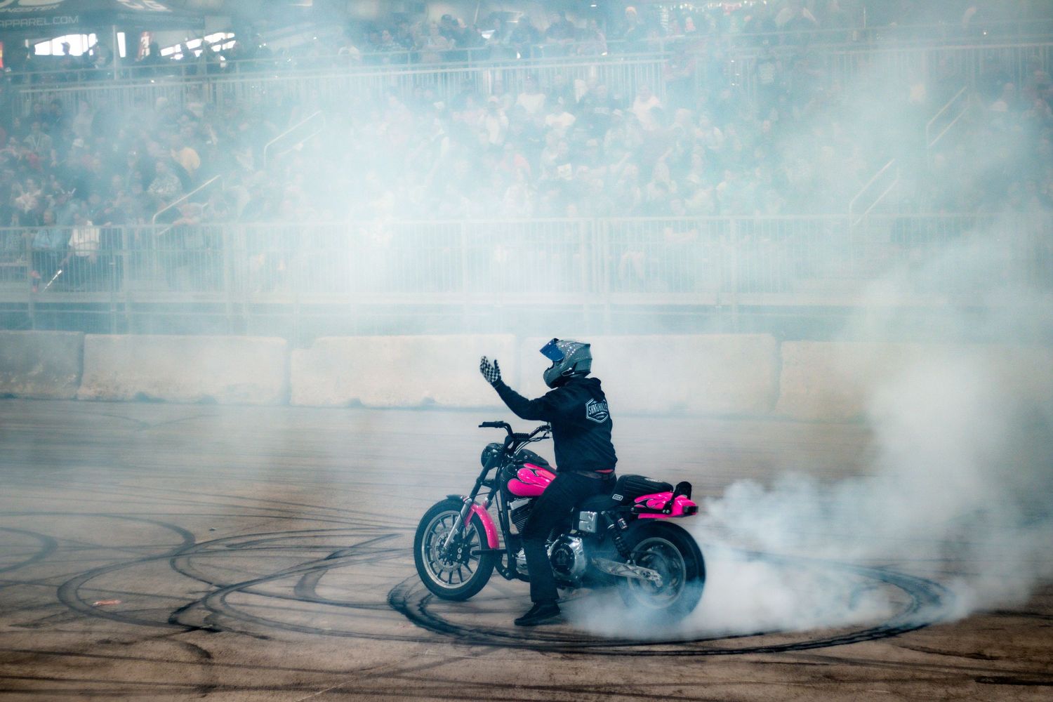 Motorcyclist performing a smoky burnout on a pink bike at an arena stunt show with crowd in background.