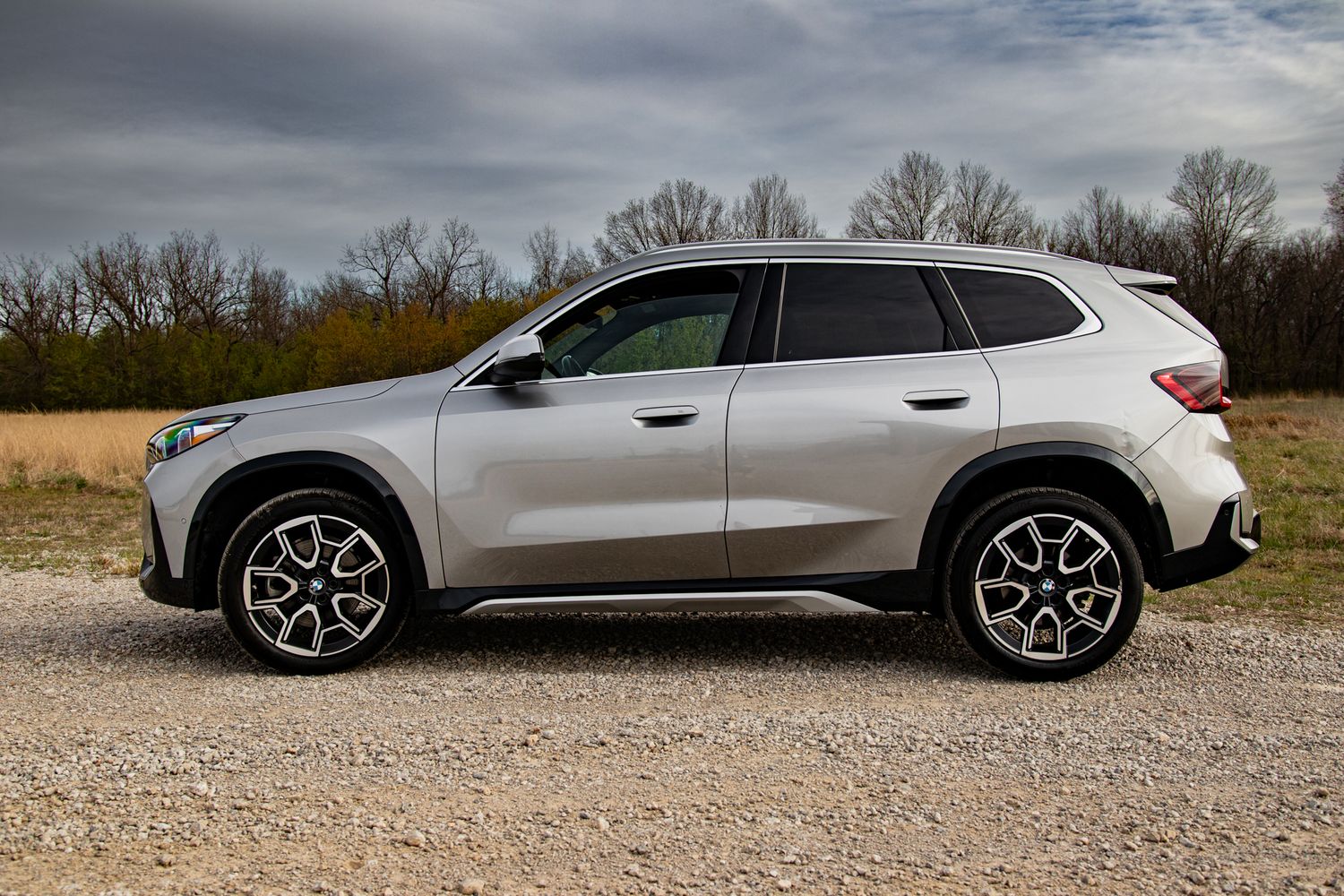 Silver SUV parked on gravel path, side profile view, stylish alloy wheels, overcast sky, trees in background.