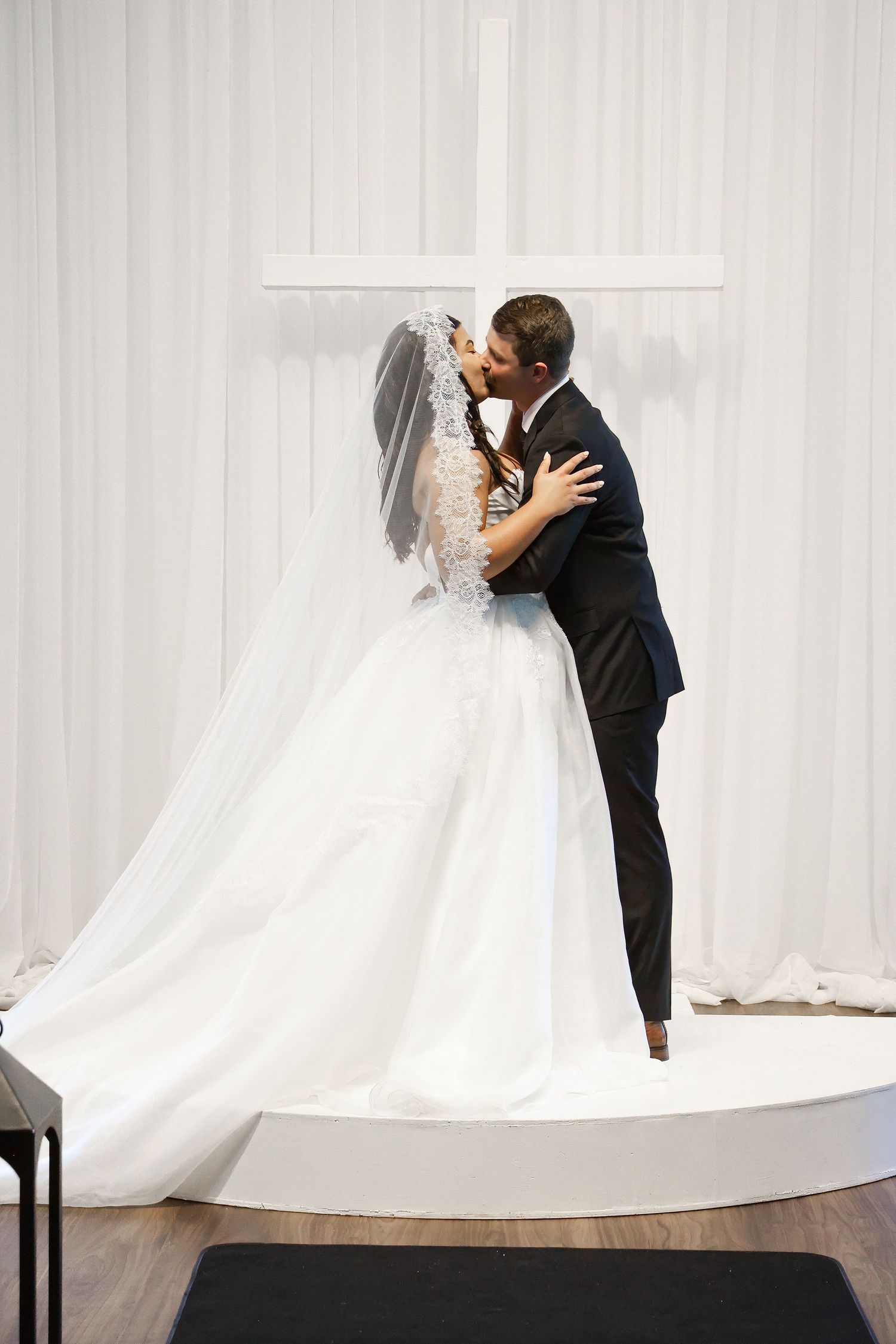Bride and groom share first kiss at altar with white cross backdrop during elegant wedding ceremony.