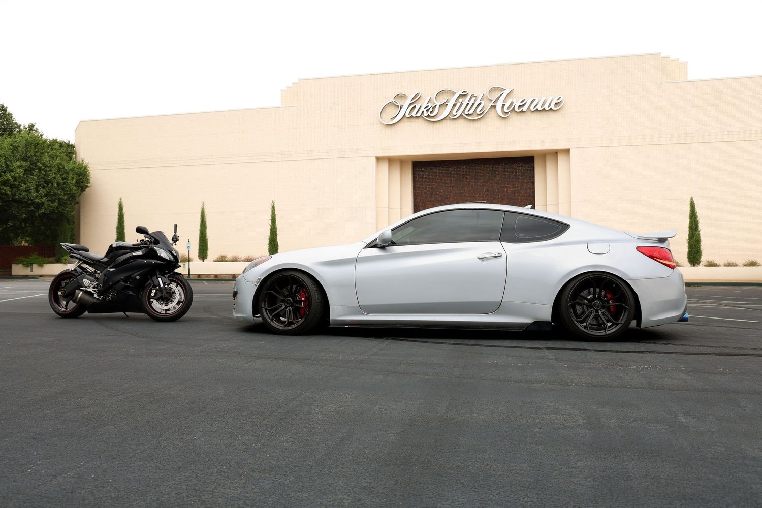 Silver sports coupe and black motorcycle parked side by side outside Saks Fifth Avenue store.