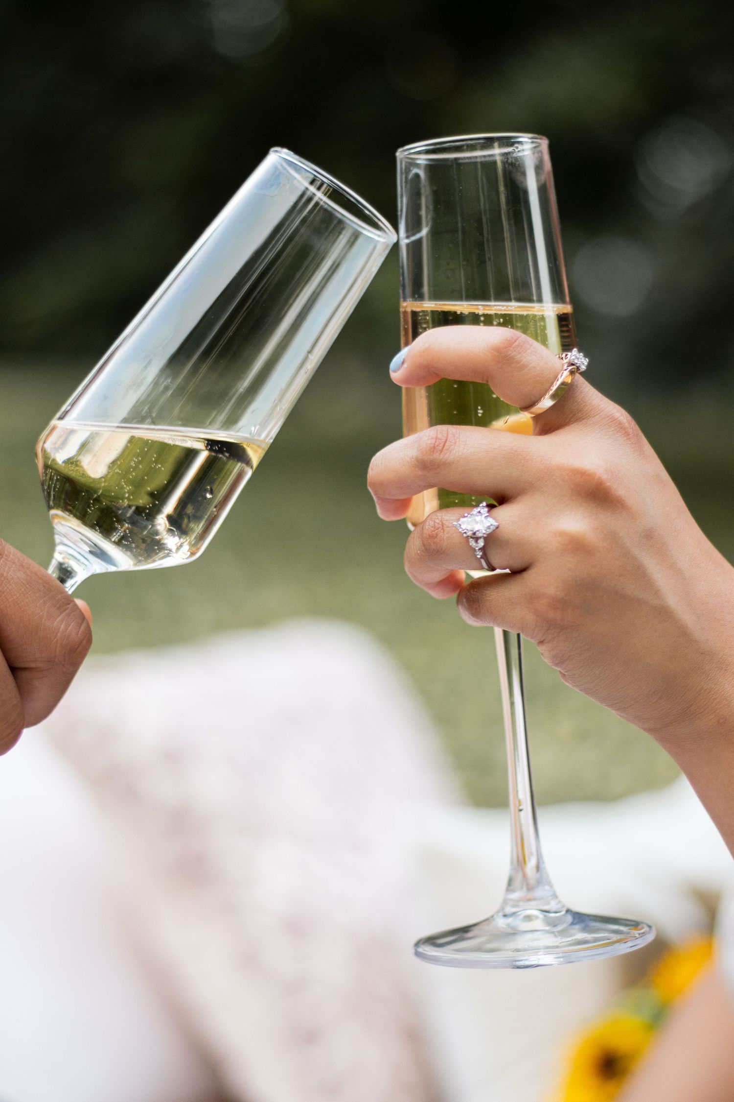 Two hands clinking champagne flutes outdoors, with a diamond engagement ring visible on one hand.