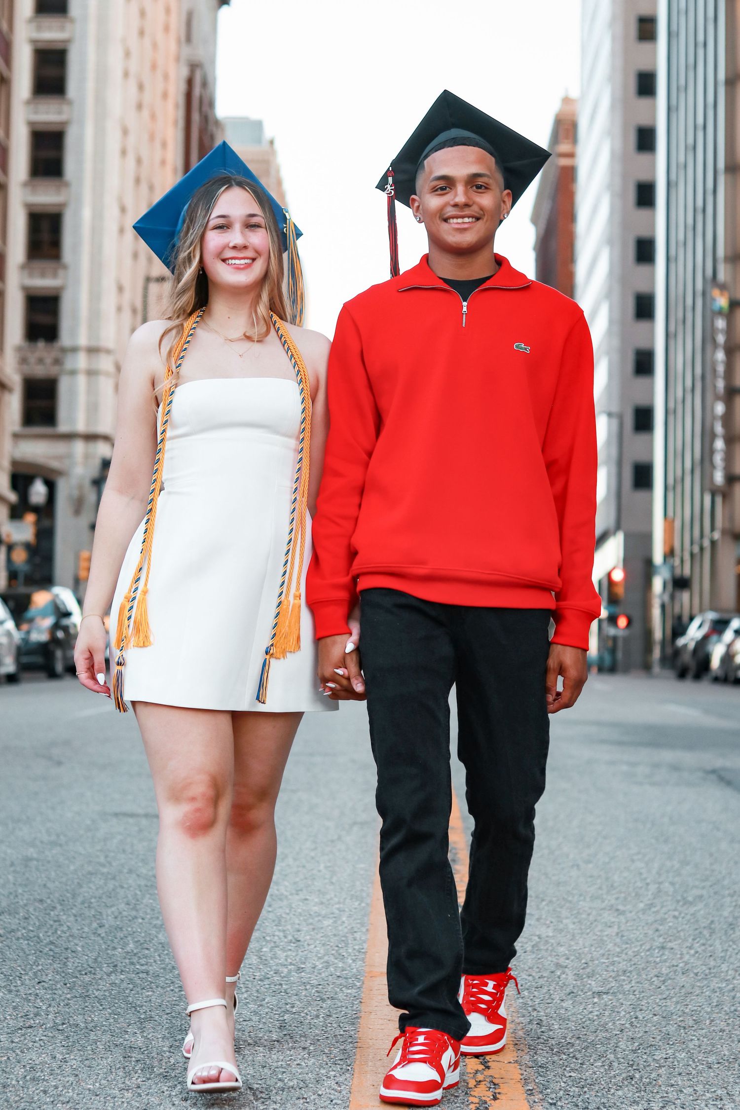 Two graduates in contrasting white dress and red hoodie walk together on a city street during commencement.
