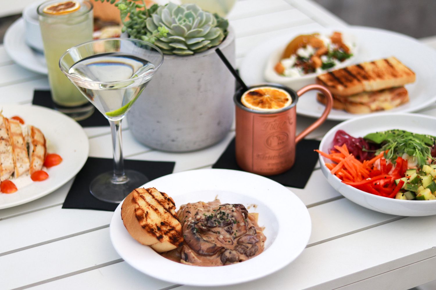 A spread of gourmet dishes including grilled meat, fresh salad, and a martini on a white outdoor dining table.