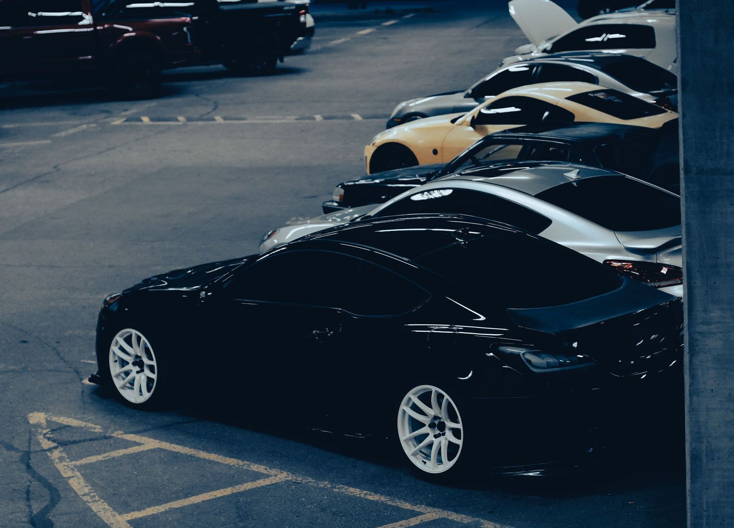 A sleek black sports car with white rims parked in a lot alongside yellow and black vehicles.