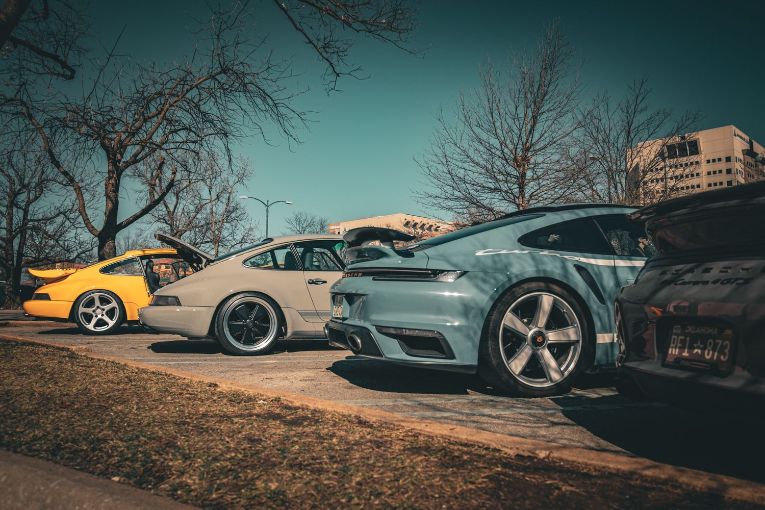 Modified sports cars including a silver Porsche and yellow car parked outdoors at a car meet on a sunny day.
