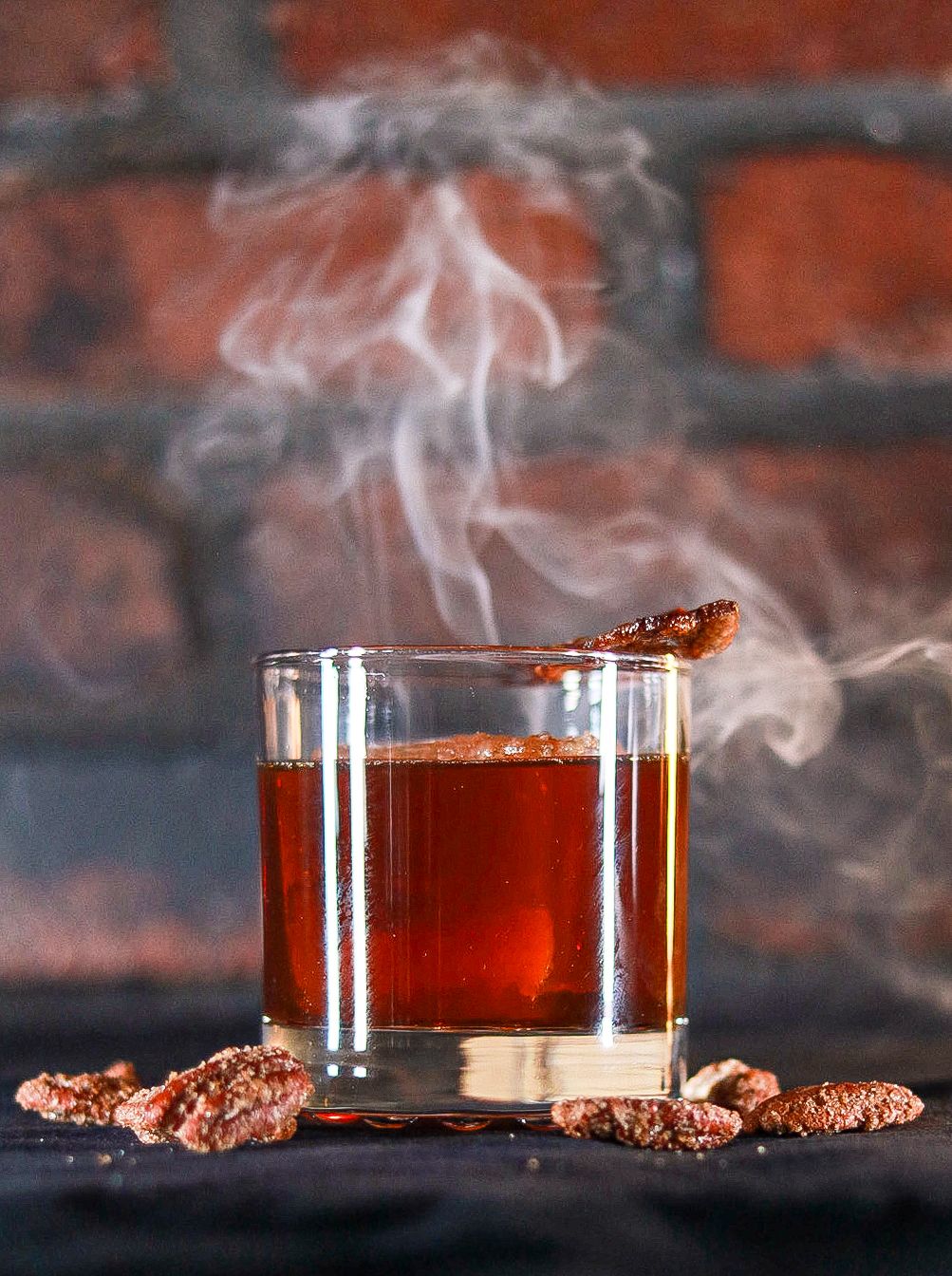 Smoky whiskey cocktail in rocks glass with candied pecans garnish on dark surface against brick wall backdrop.