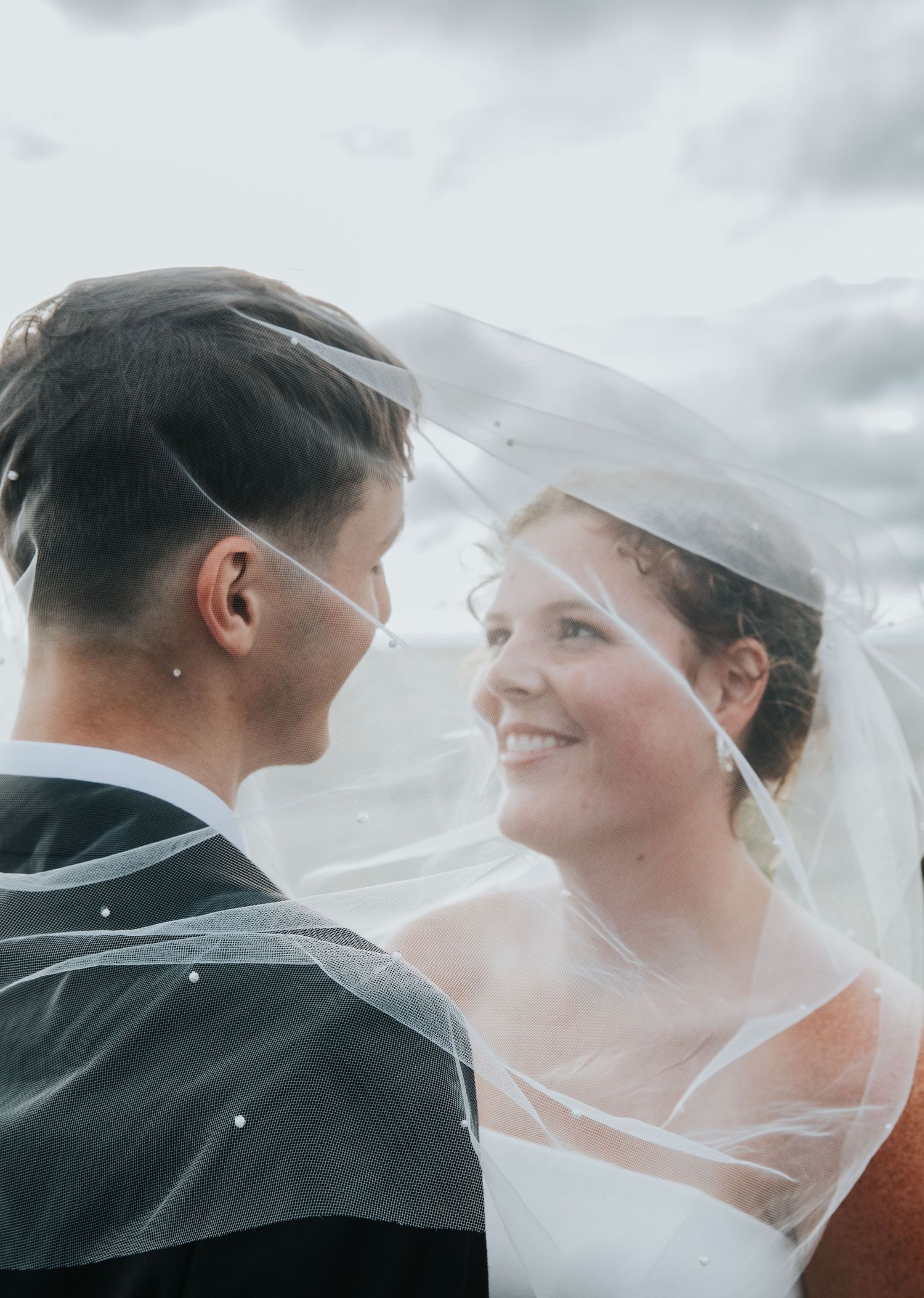 A smiling bride in a flowing veil gazes at her groom on their wedding day under a cloudy sky.
