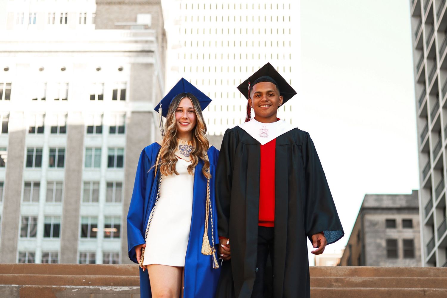 Two graduates in academic regalia stand together on steps against an urban backdrop with tall buildings.