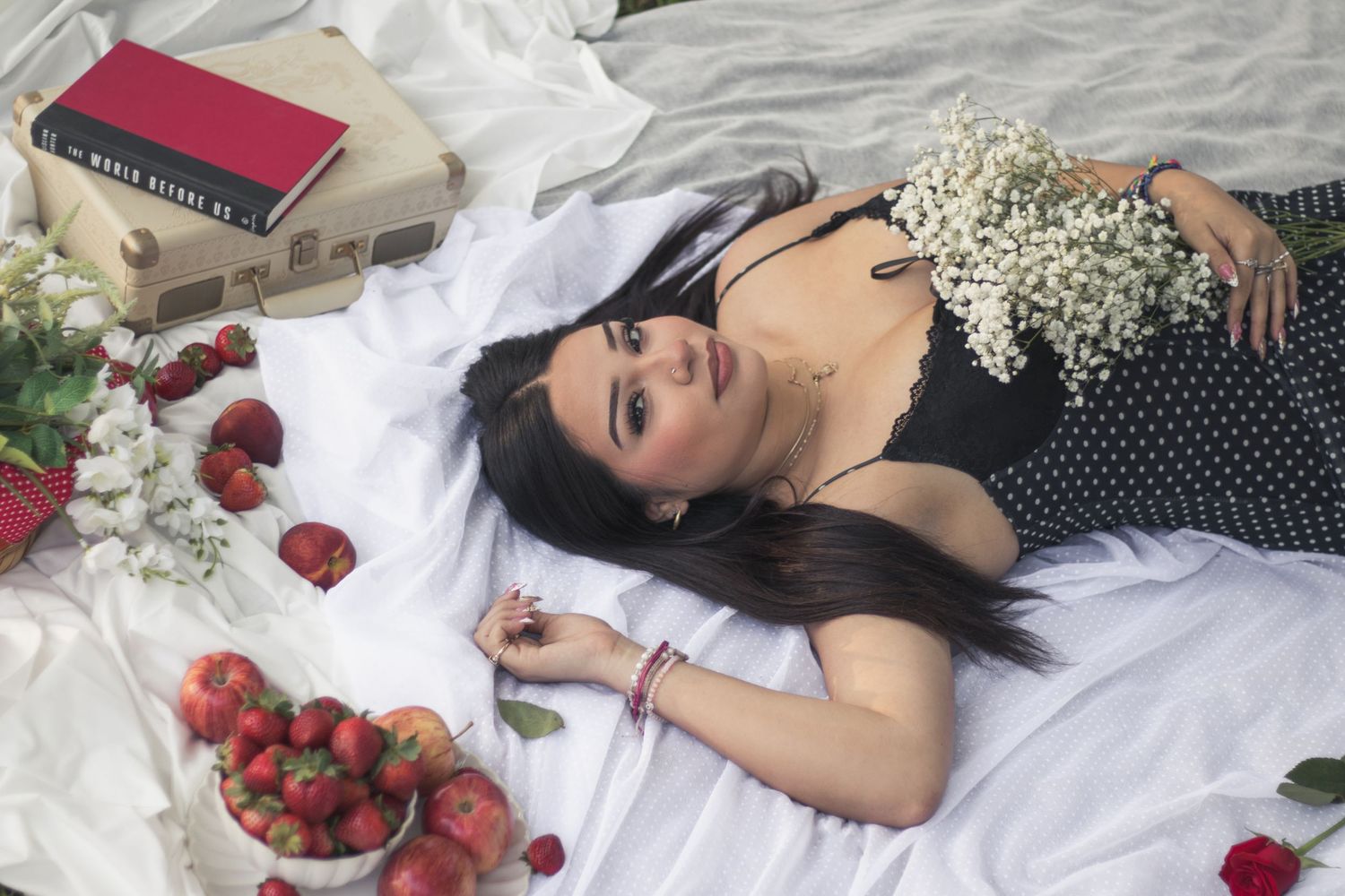 Woman lying on white bed surrounded by strawberries, roses, and white flowers, wearing a black polka dot dress.