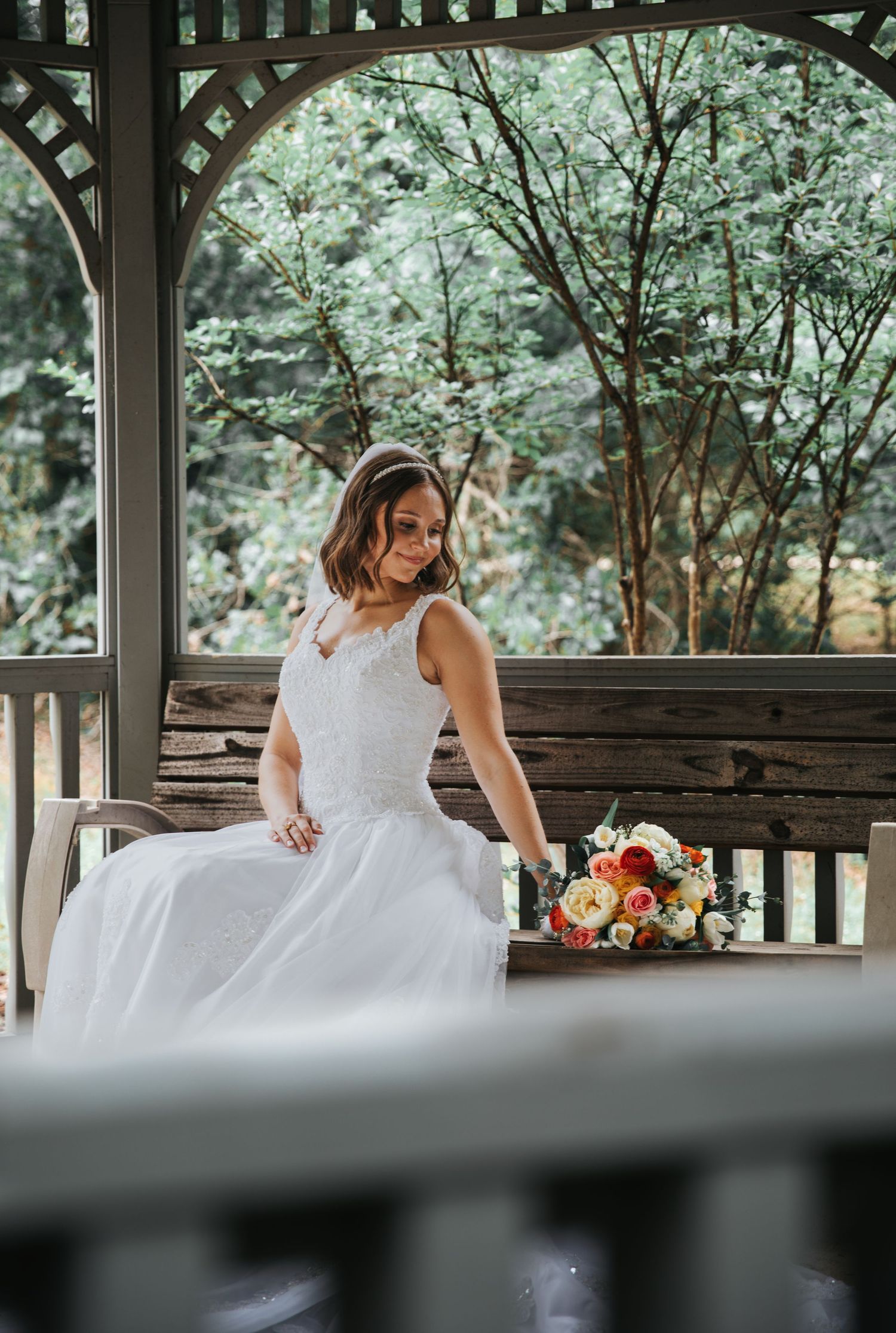 Bride in white lace wedding gown sitting on bench in gazebo with colorful bridal bouquet surrounded by lush greenery.