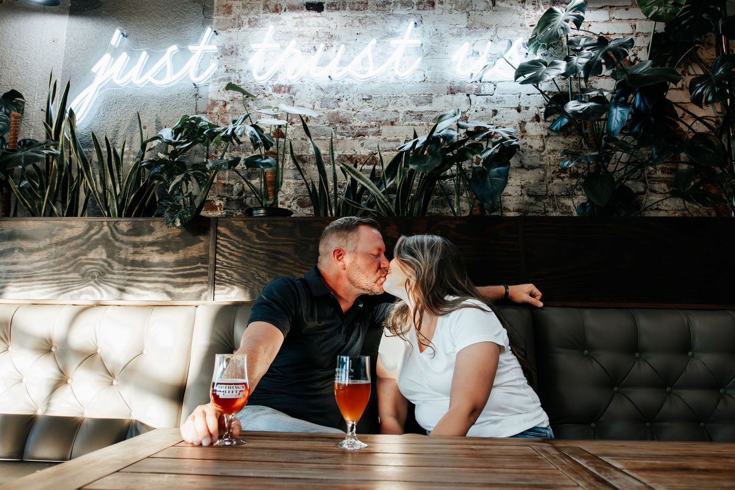 Couple kissing at a restaurant booth with craft beers and a neon 'just trust' sign on a brick wall.