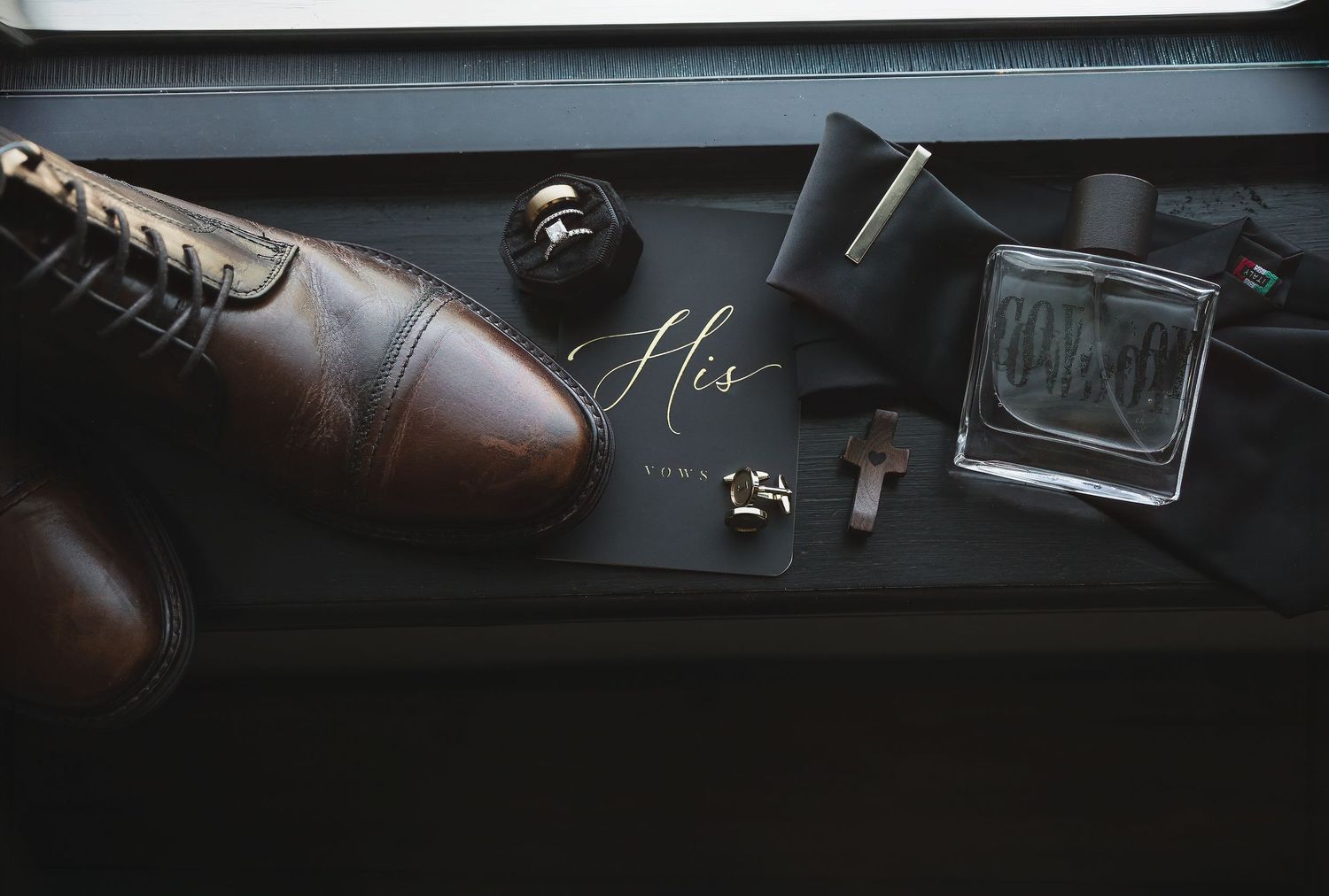 Groom wedding flat lay with brown leather dress shoes, cologne, cufflinks, bow tie, and a black His card.