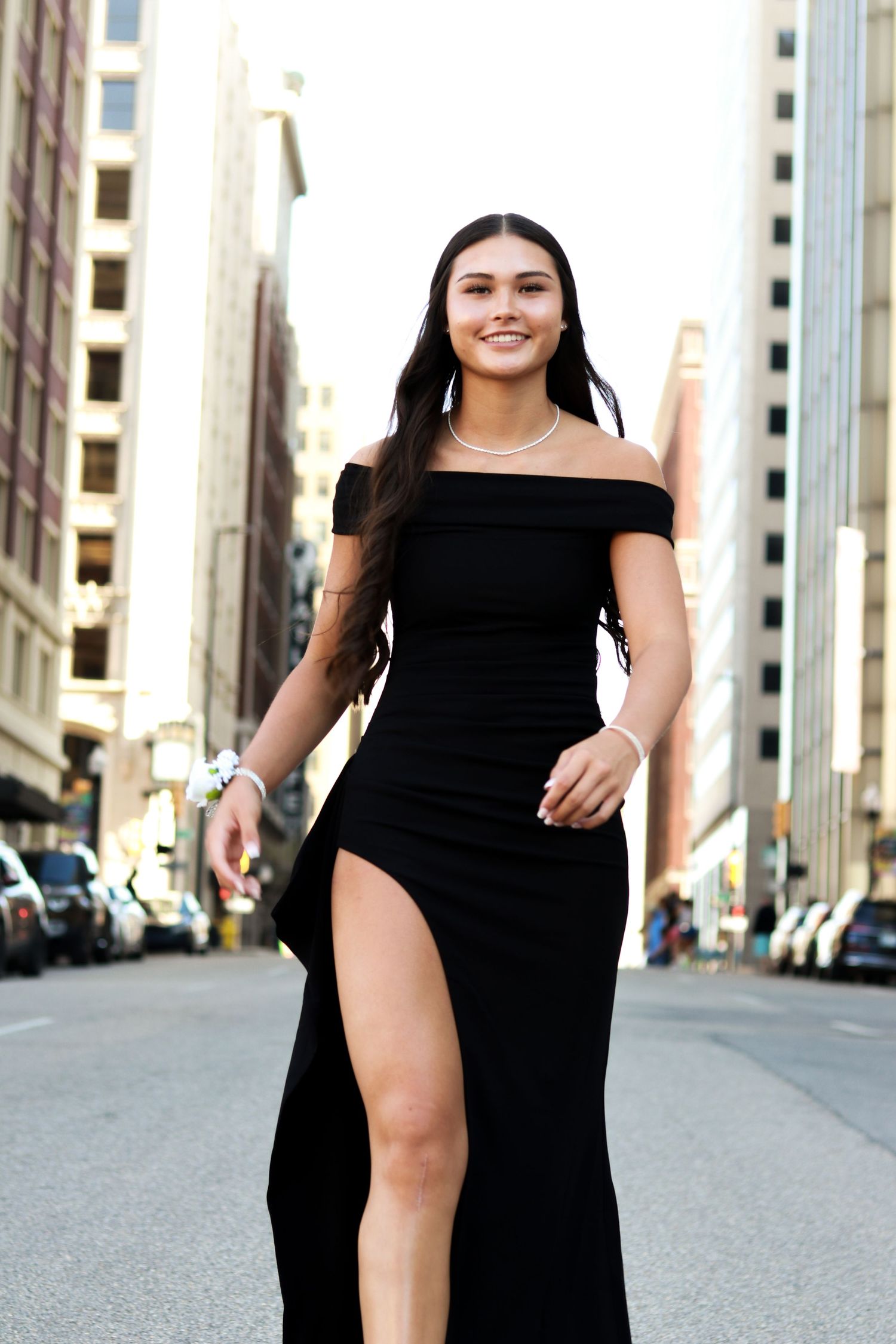 A person in a black dress poses confidently on an urban street with tall buildings in the background.