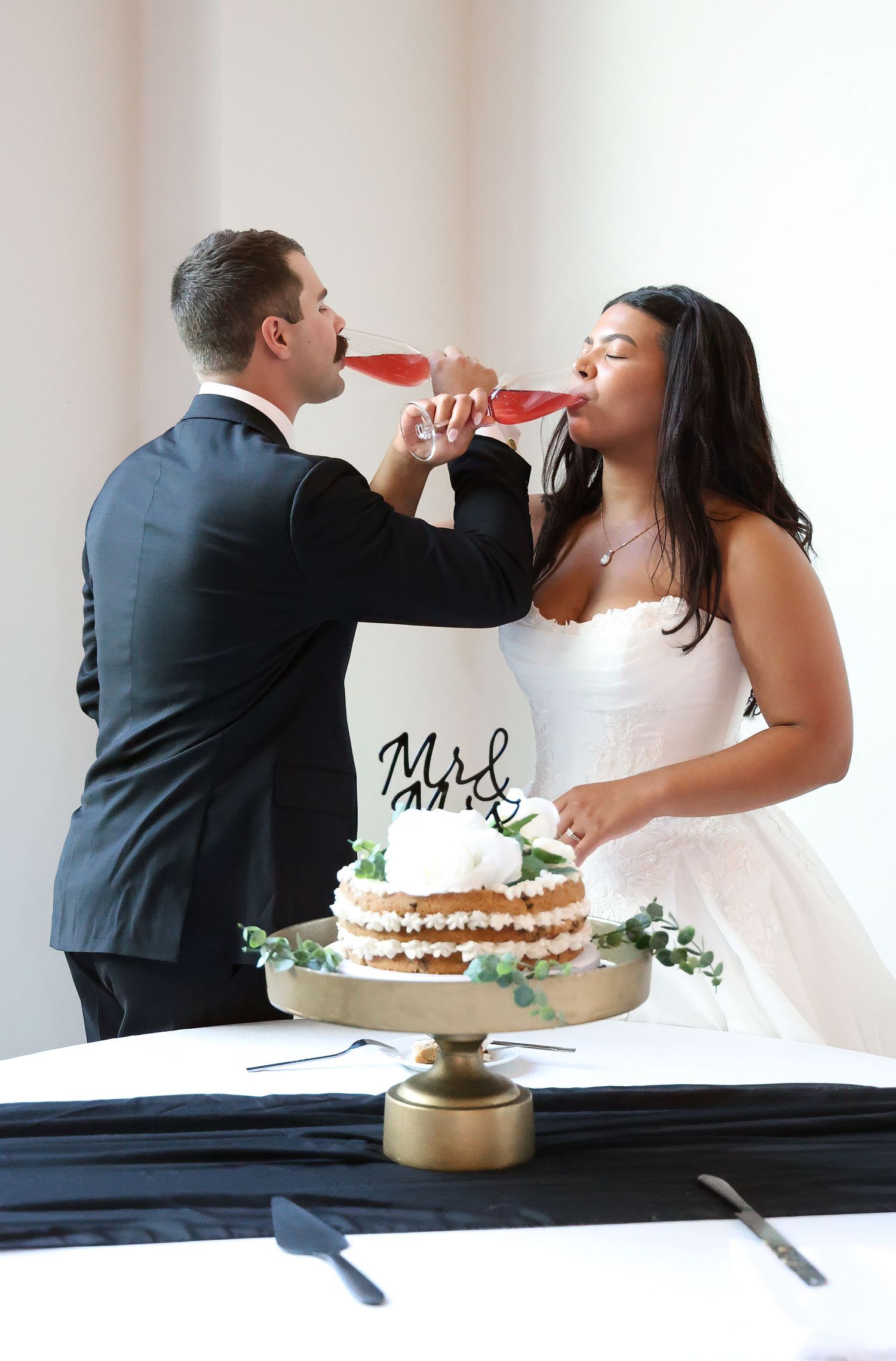 Bride and groom sharing a drink from a wine bottle beside their wedding cake with Mr & Mrs topper.