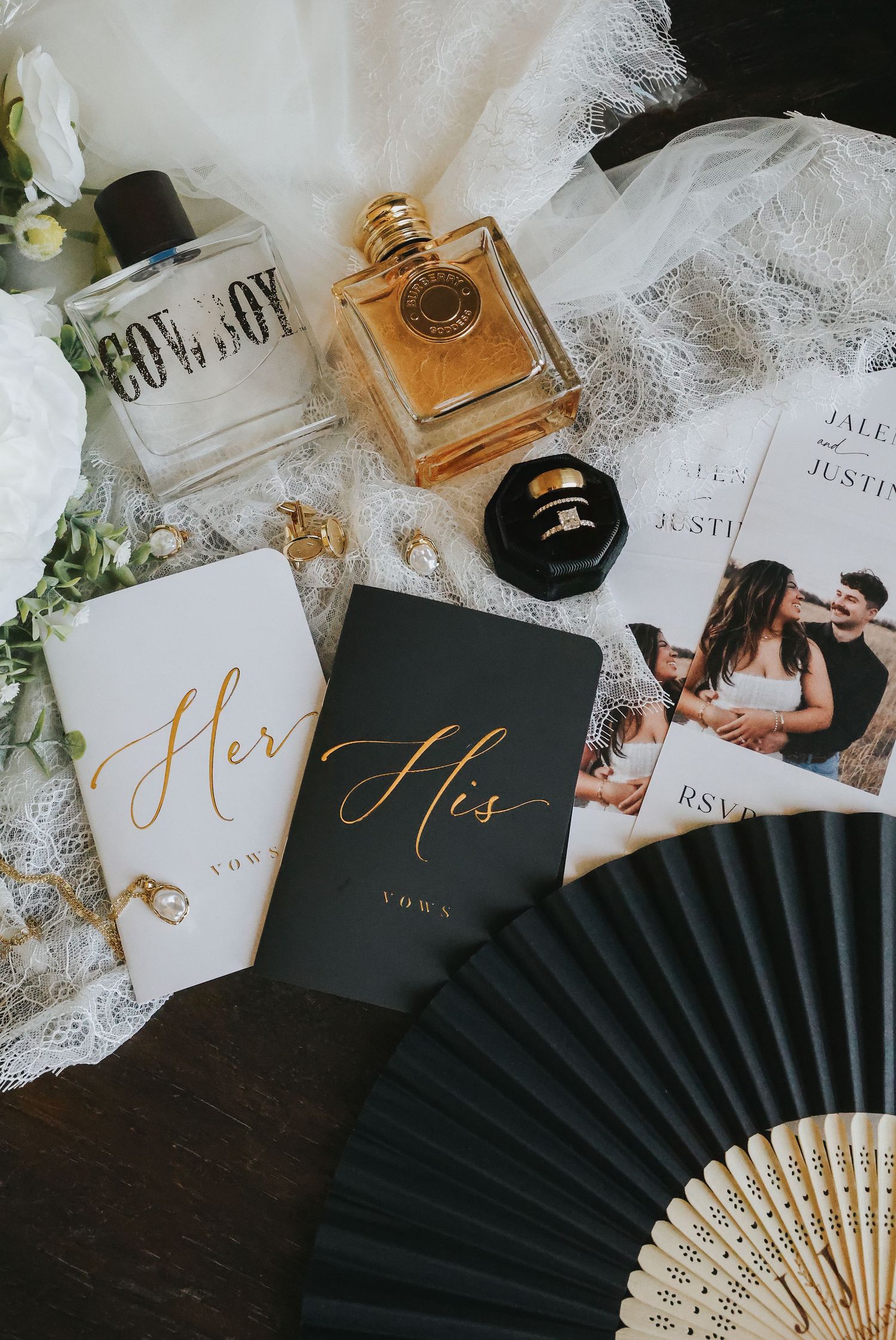 Elegant wedding flat lay with His and Her vow books, perfume bottles, rings, invitations, and a black fan on lace.