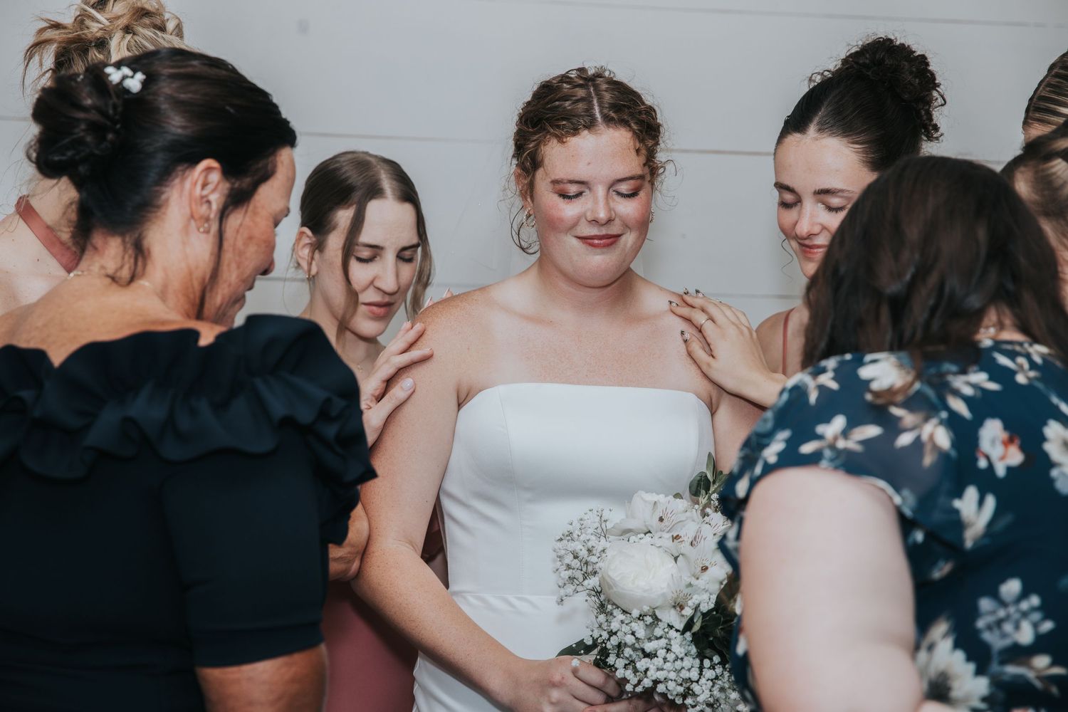 Bride in white strapless gown holding bouquet surrounded by bridesmaids in black dresses at wedding.