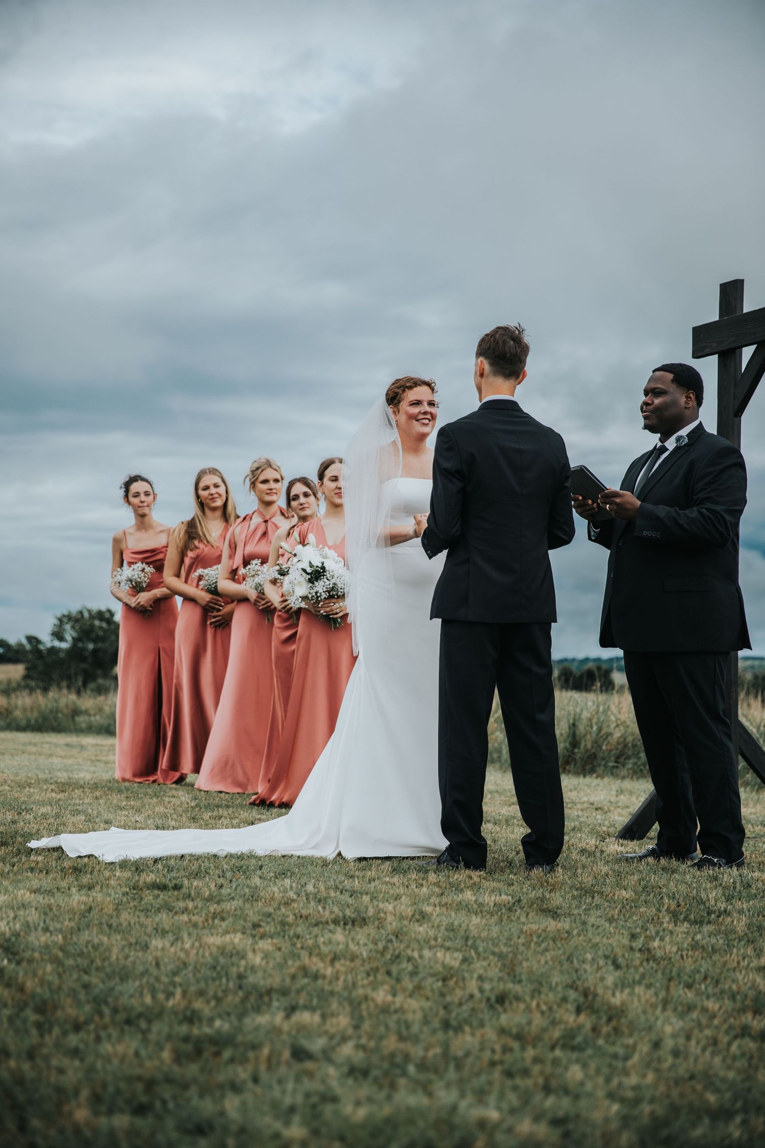 Outdoor wedding ceremony with bride in white gown, bridesmaids in coral dresses, groom in black suit, and officiant holding bible.