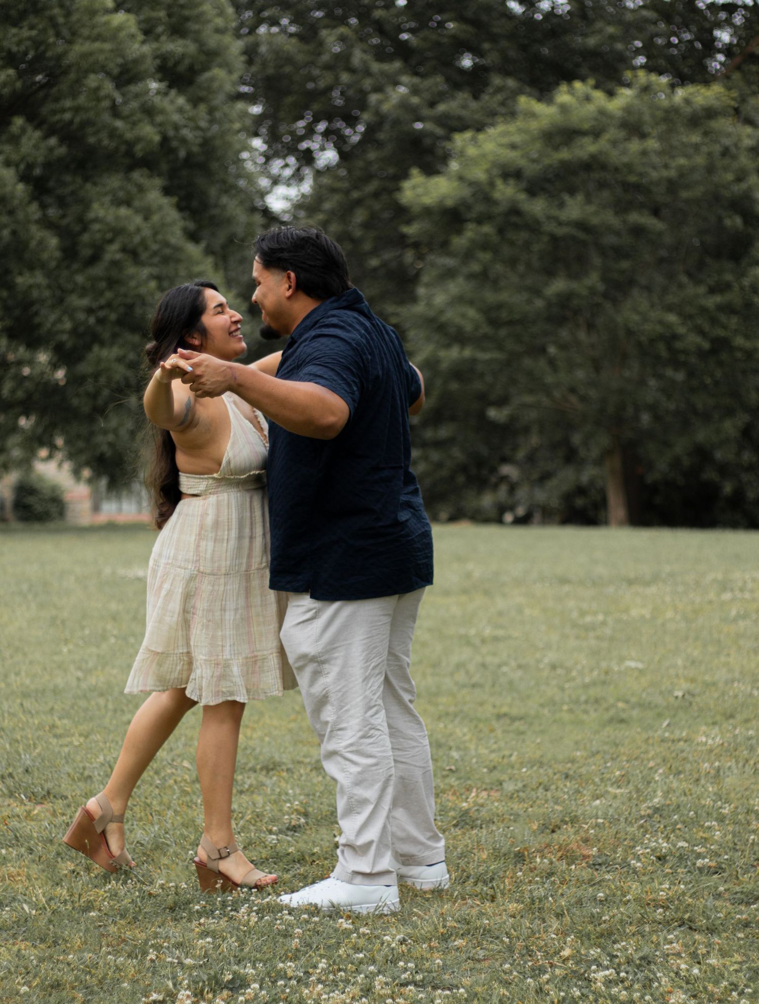 A joyful couple dancing and laughing together in a lush green park, woman in white dress, man in navy shirt.
