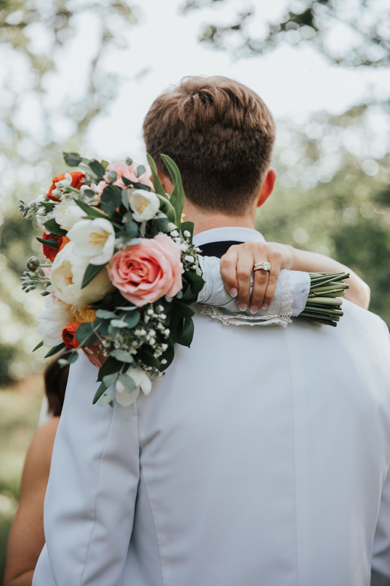 Bride with wedding bouquet of pink and white roses embracing groom in white suit from behind outdoors.