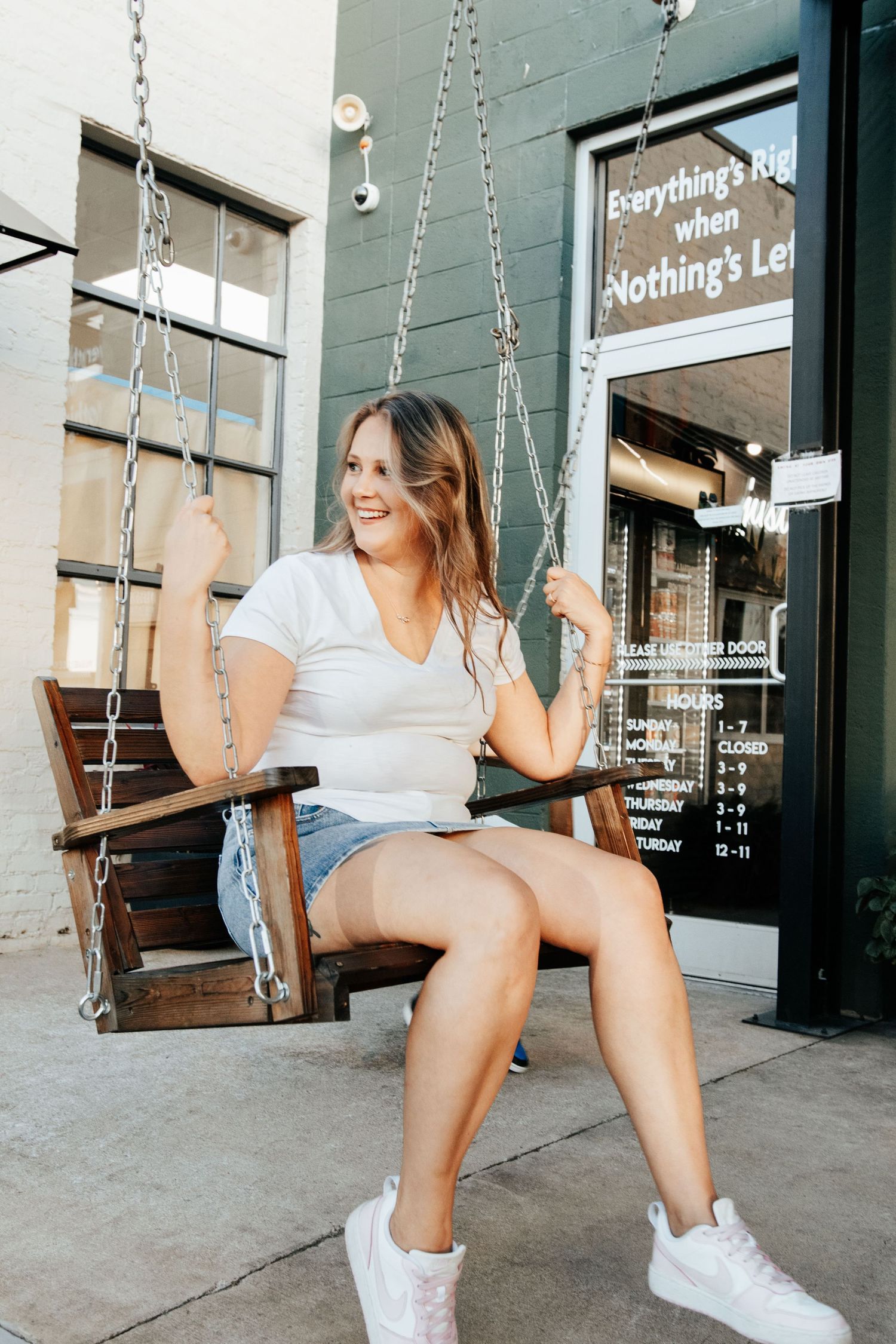 Young woman in white tee and denim shorts smiling on a wooden chain swing outside Nothing's Left Brewing Co.