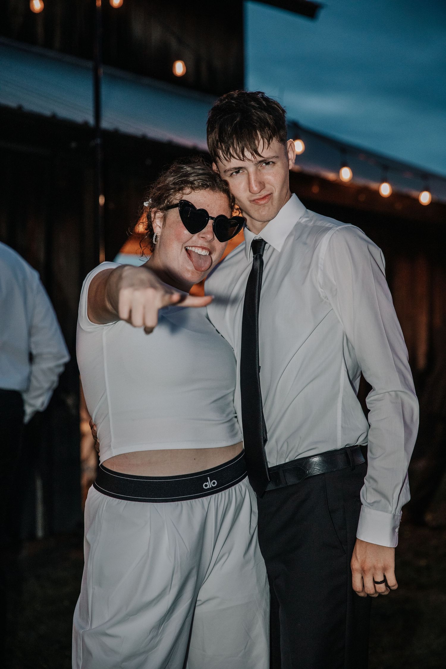 Two young people pose together at an outdoor evening event, woman in white crop top with heart sunglasses pointing at camera.