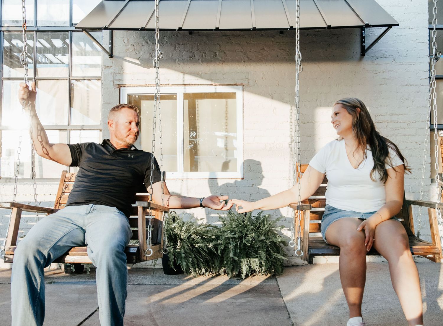 A man and woman sit on wooden porch swings, smiling and reaching toward each other outdoors.