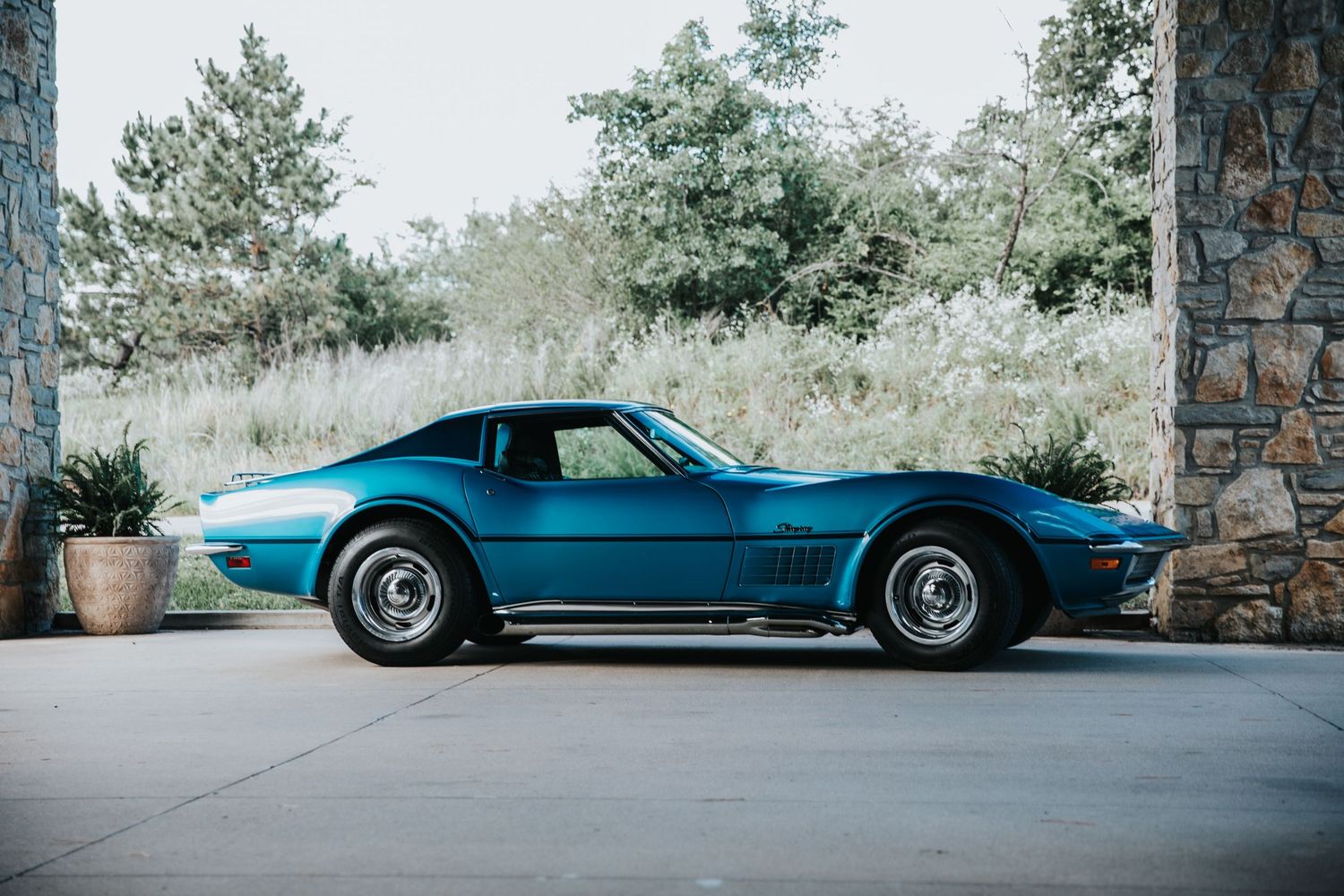 Classic blue 1969 Chevrolet Corvette Stingray parked on concrete driveway beside stone wall and lush green trees.