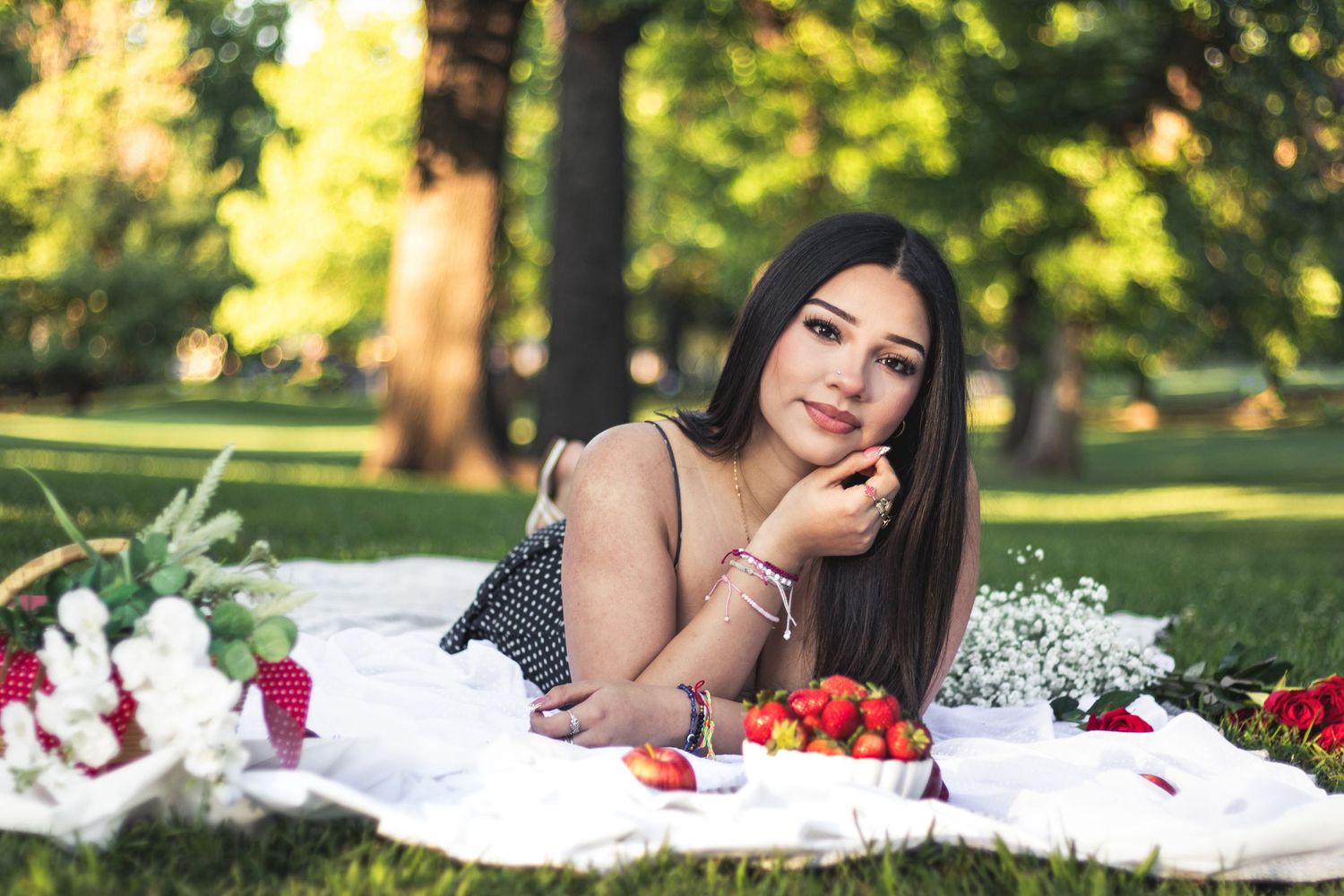Young woman lying on a white blanket in a park, enjoying a picnic with strawberries and flowers on a sunny day.