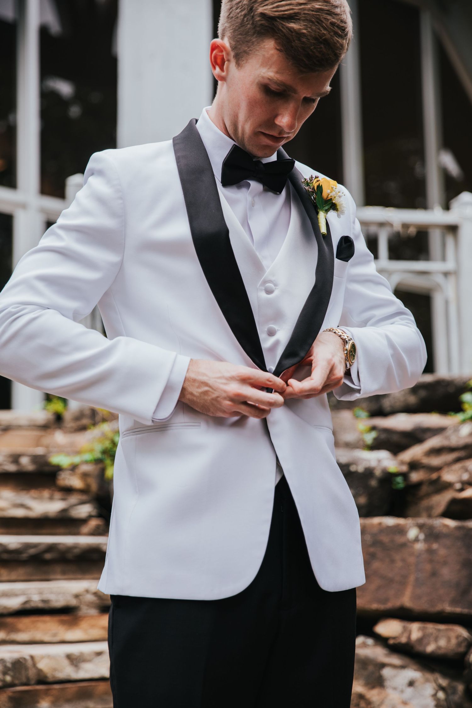 A groom in a white tuxedo jacket with black lapels and bow tie buttons his jacket outdoors before his wedding.