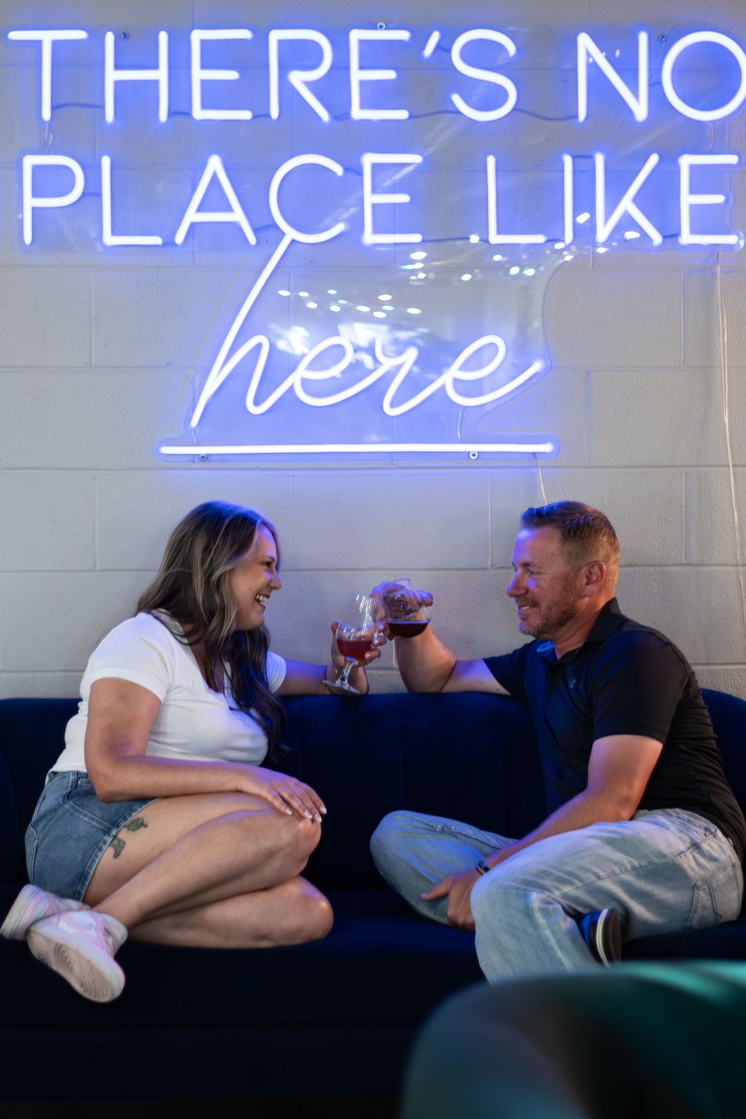 Couple toasting wine glasses on blue sofa under neon sign reading There's No Place Like Here.