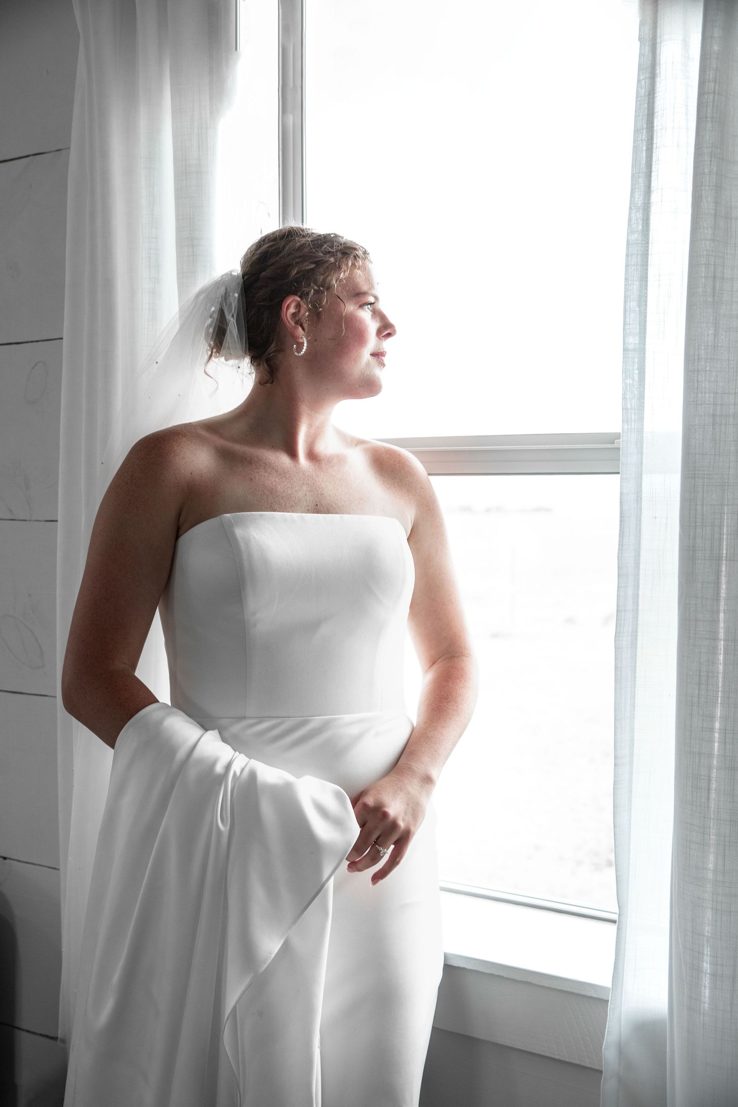 Elegant bride in strapless white gown and veil gazing thoughtfully out a bright window on her wedding day.