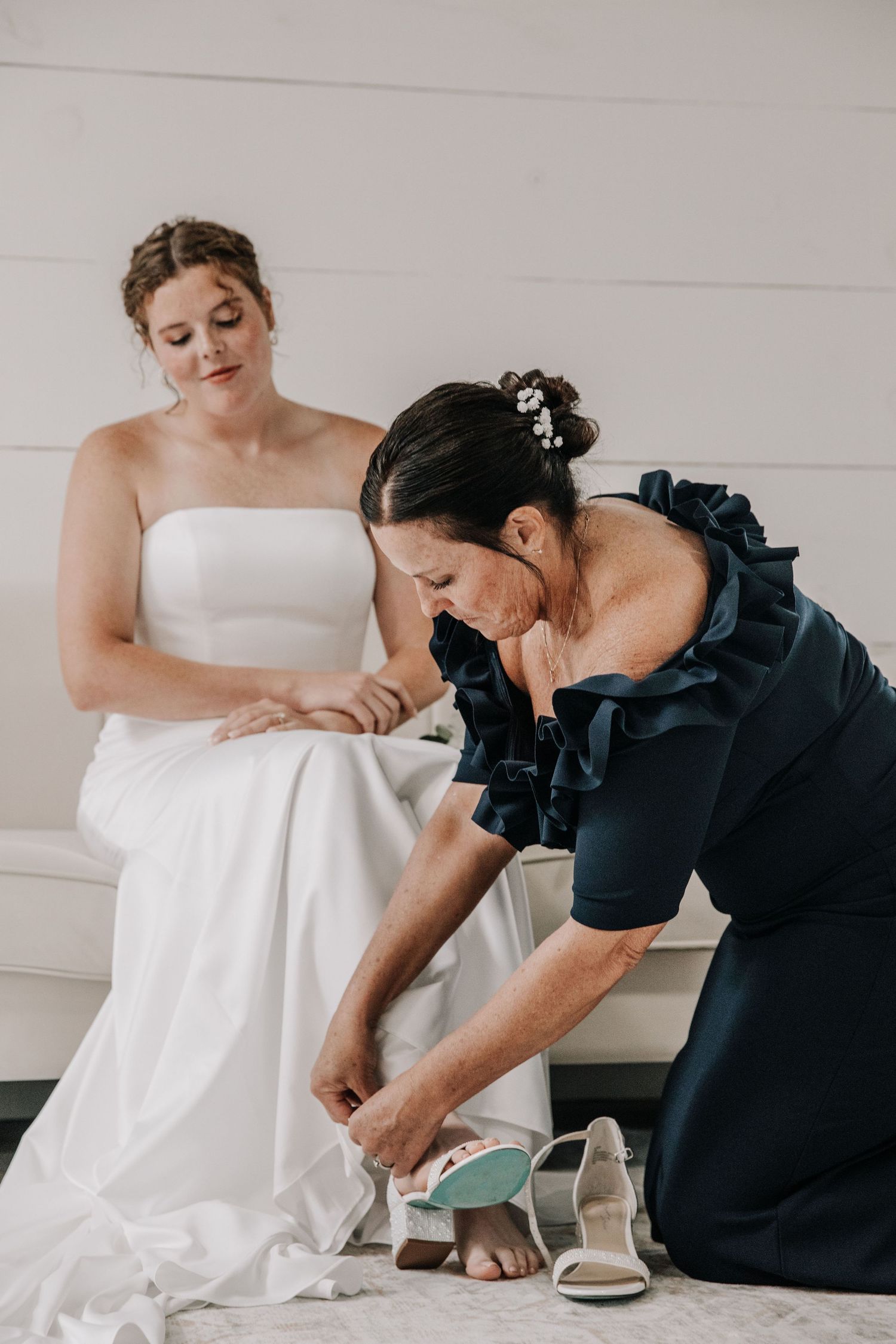 A bride in a white strapless gown sits as a woman in navy blue helps her put on wedding shoes.