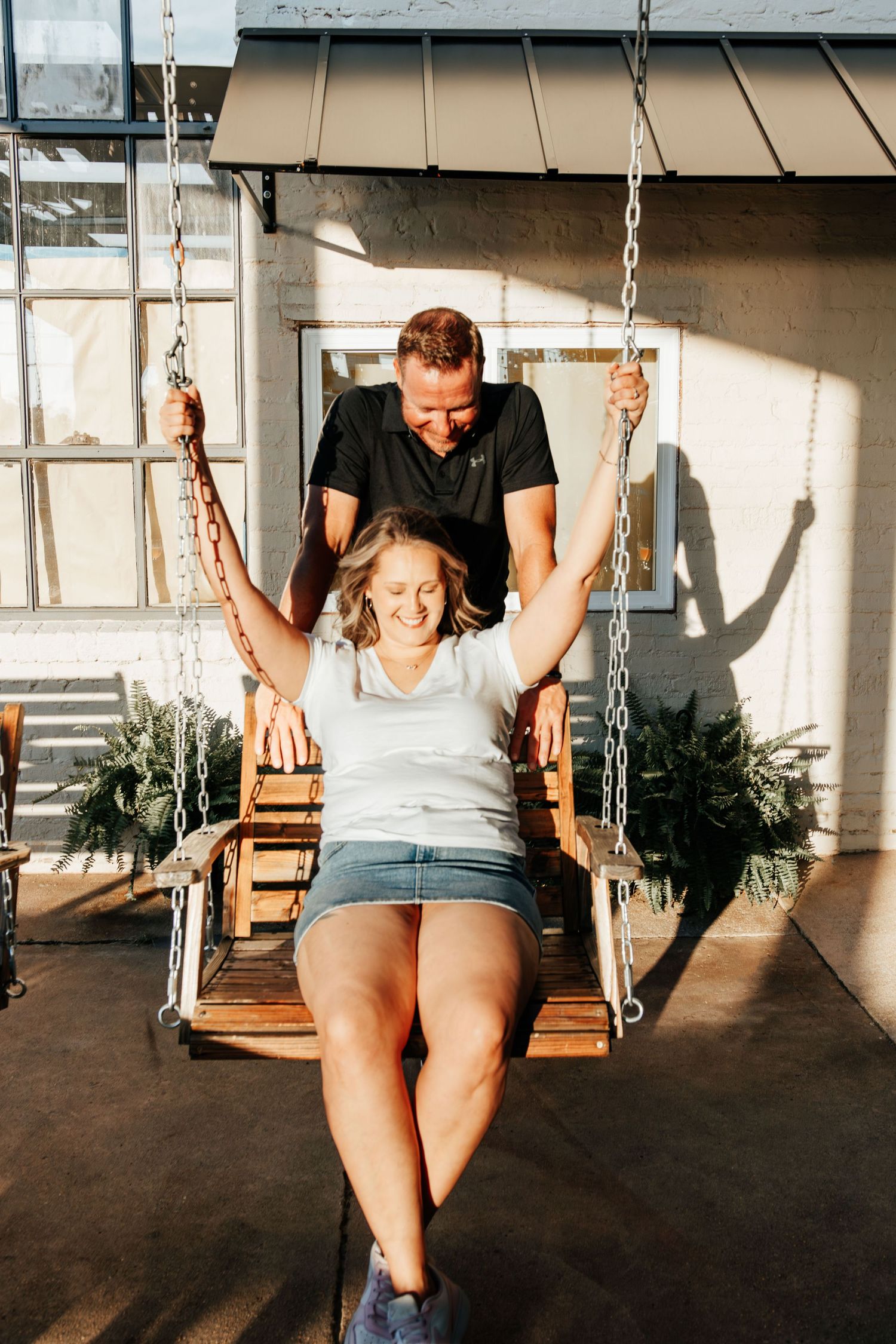 Man pushes smiling woman on wooden porch swing in sunny outdoor setting.