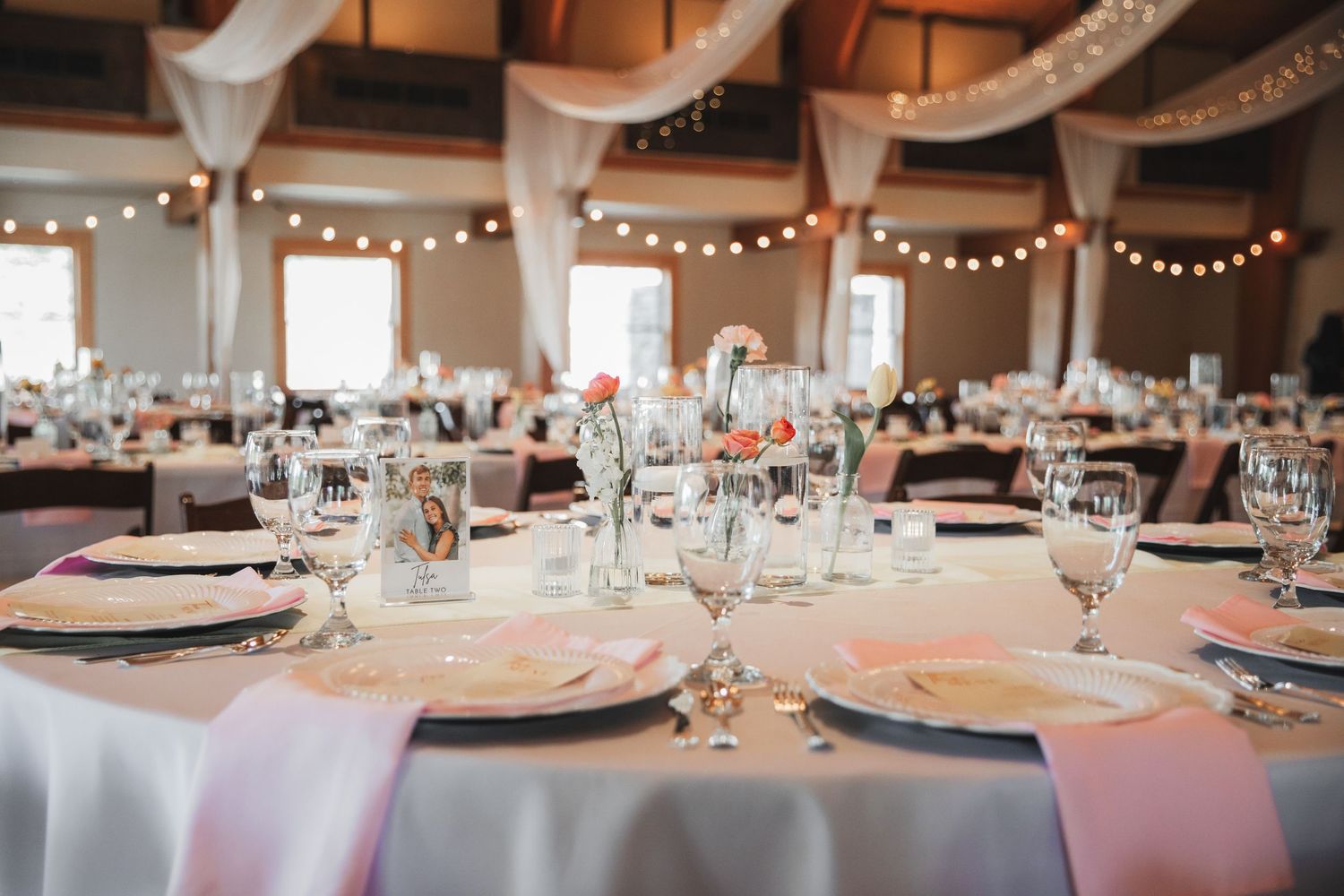Elegant wedding reception hall with pink satin napkins, crystal glassware, floral centerpieces, and draped white fabric ceiling decor.